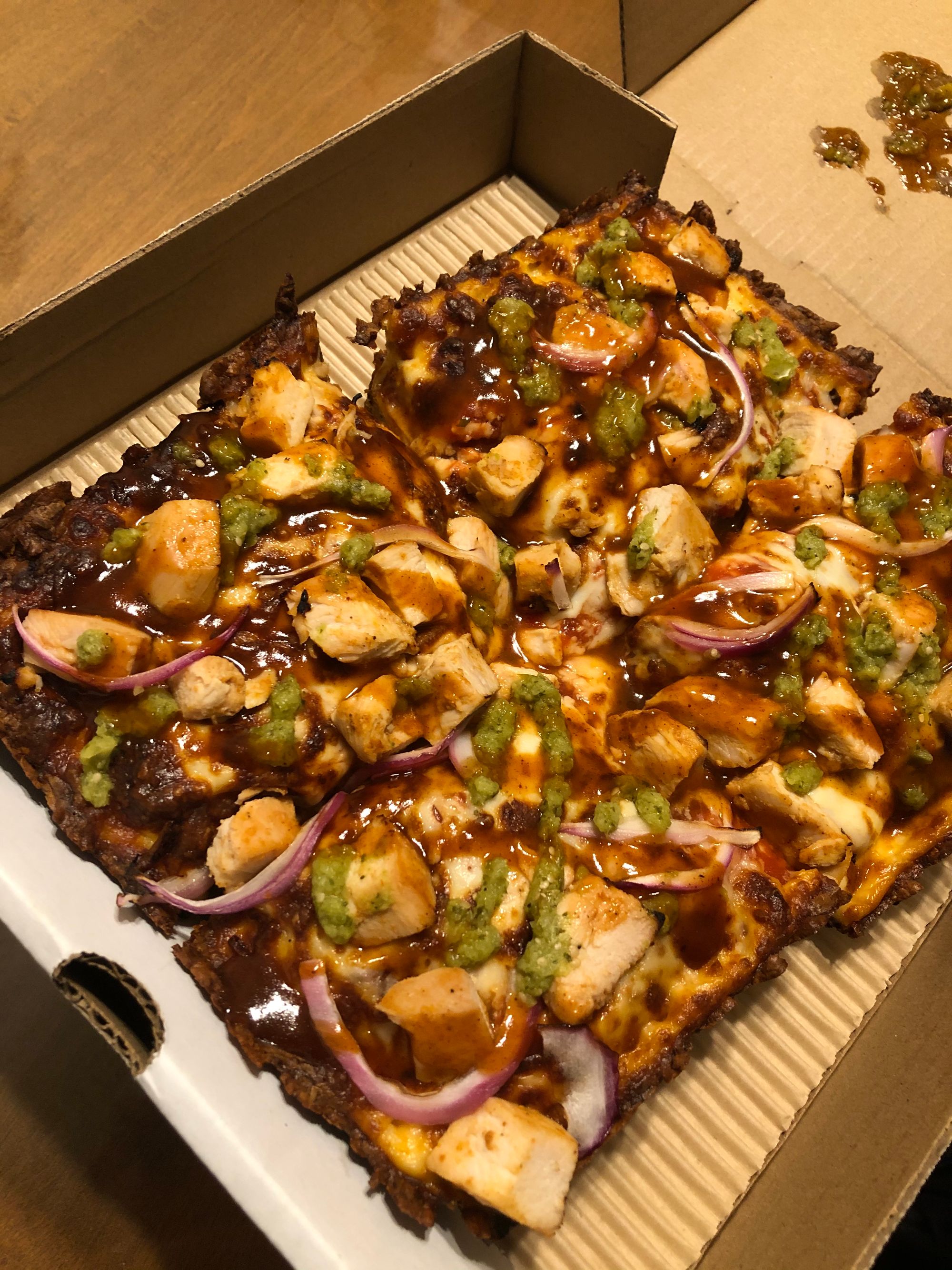 A Detroit-style pizza: square, in a cardboard box, covered in chunks of chicken, bbq sauce, salsa verde and red onions.