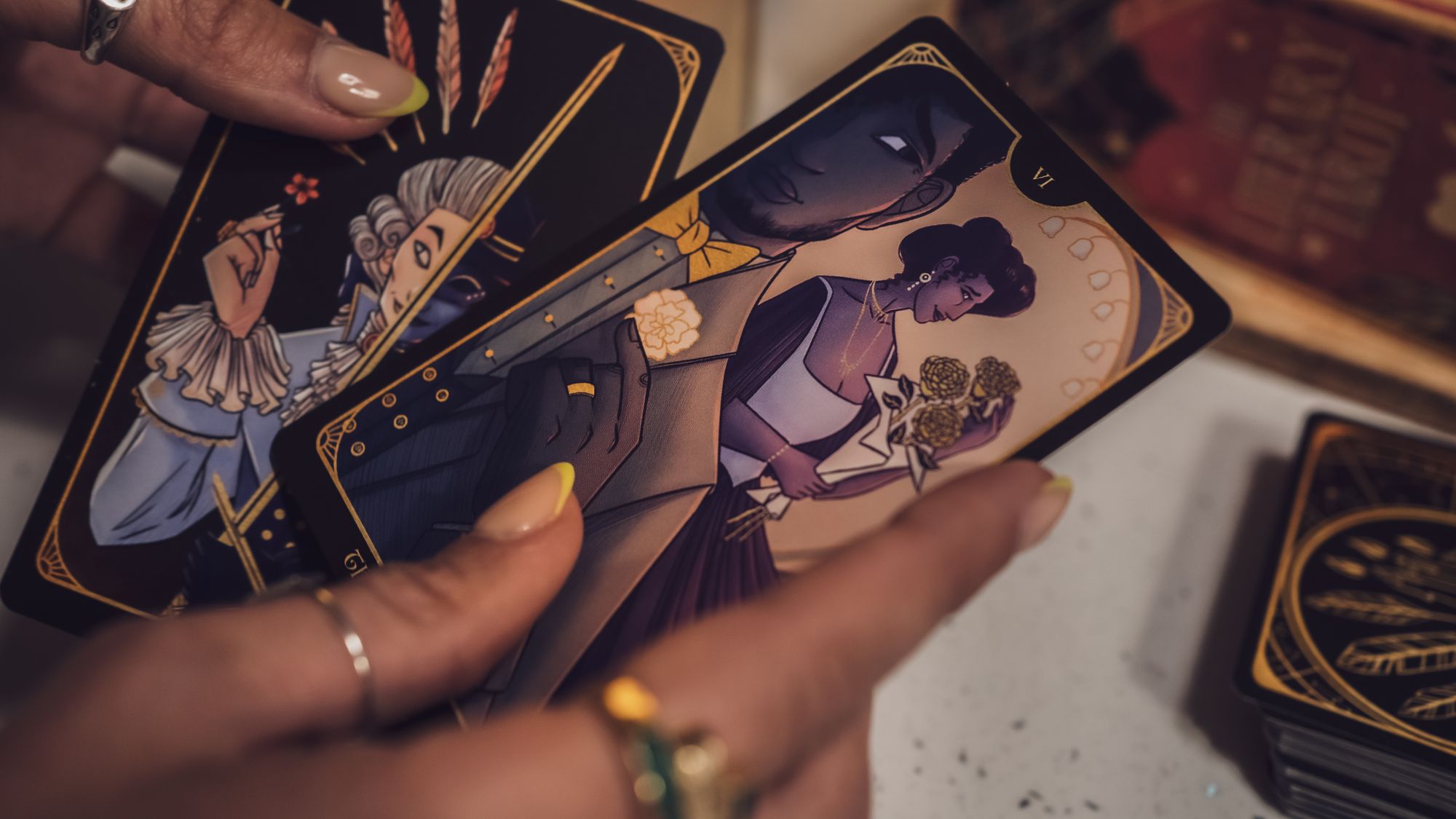 A pair of dark-skinned and well-manicured hands wearing rings hold two illustrated Tarot cards face up from the deck, against a blurred background of stacked, face-down cards showing off the lovely gold-on-black backs. In one face-up card (The Lovers), a Black man in a handsome waistcoat and jacket looks hopefully over his shoulder at an elegant Black woman holding a bouquet of three golden roses.