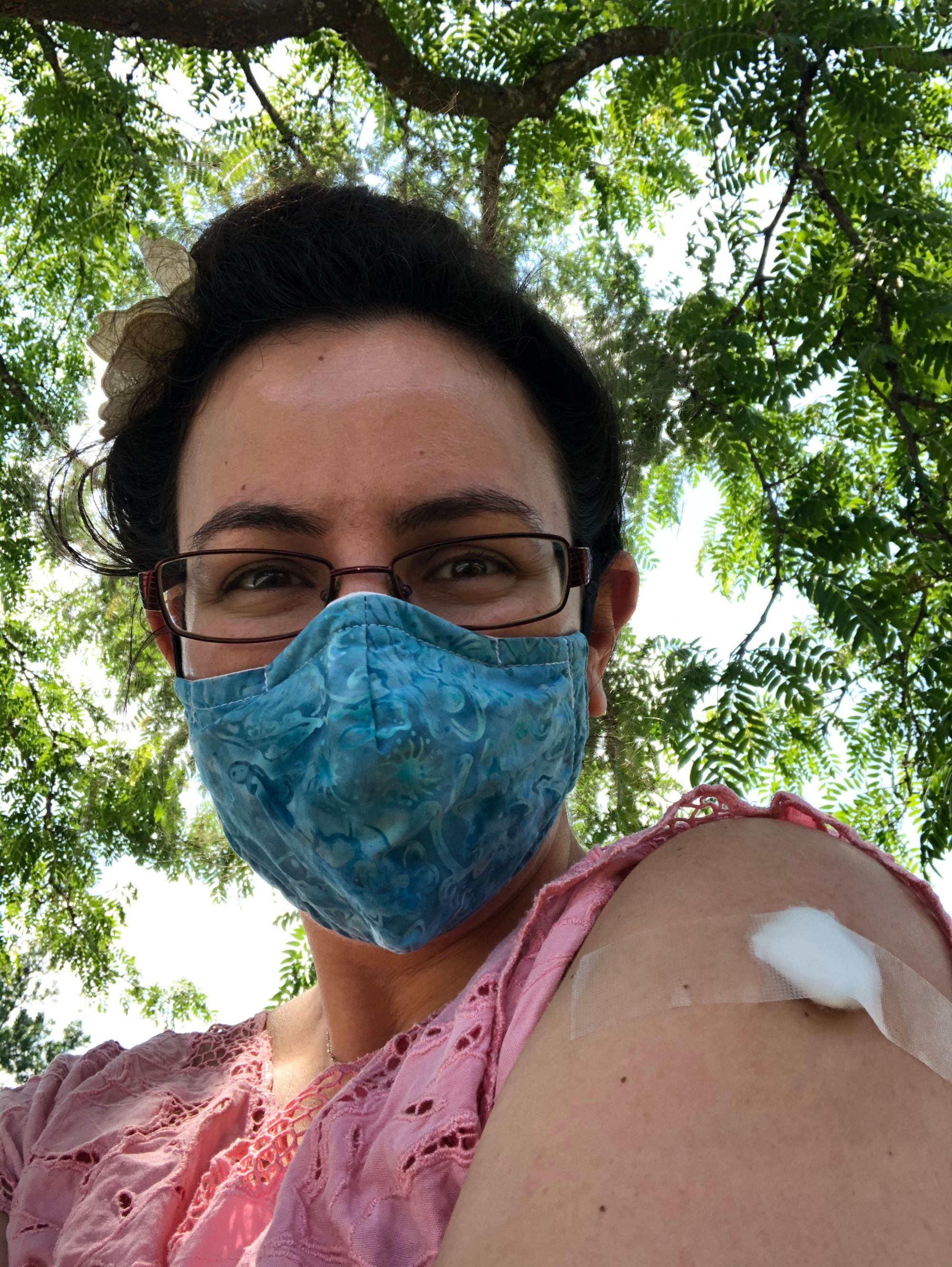Close up selfie against a sunny backdrop of green leaves; I'm wearing rectangular glasses and a turquoise mask in a damask pattern and a pink lacy shirt, showing off the taped cottonball over my vaccinated shoulder.