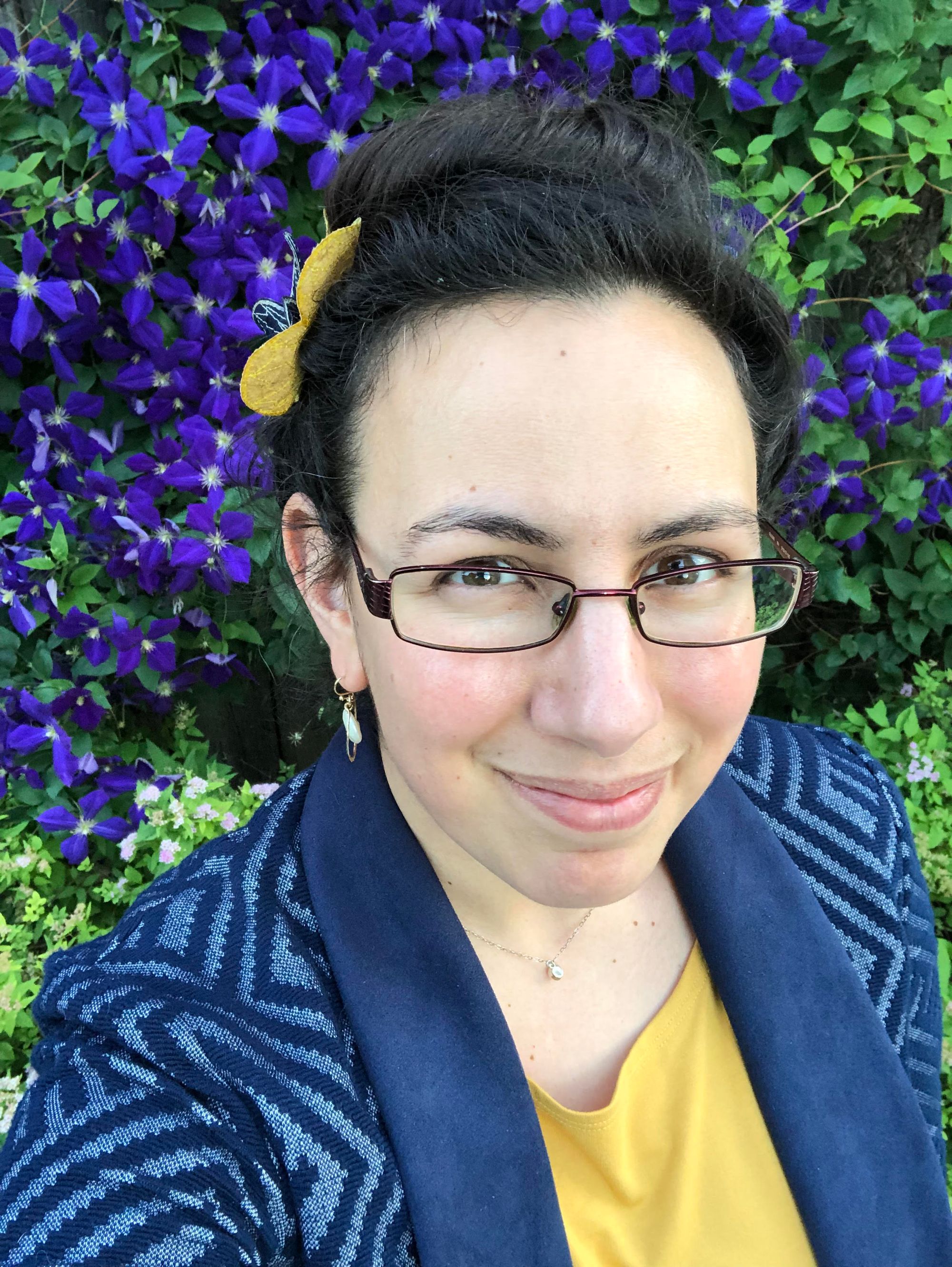 Selfie: I'm wearing glasses and a blue blazer with a geometric pattern over a bright yellow t-shirt against a backdrop of spectacularly purple-blue clematis flowers. I have two silk flowers in my pinned-up dark hair: one bright yellow and one dark blue. I'm wearing a tiny pearl necklace on a short delicate chain and larger freshwater-pearl earrings in a rose-gold setting.  