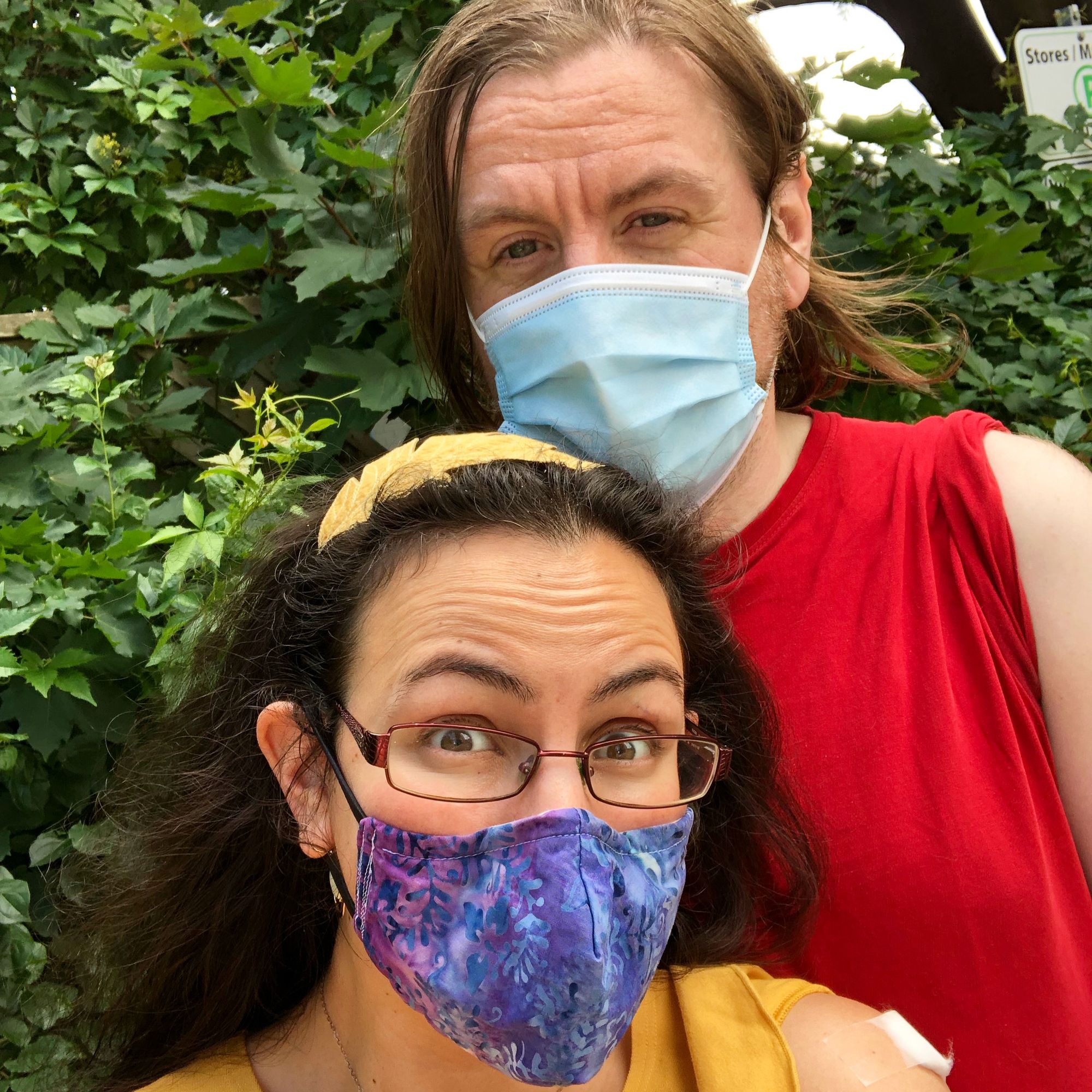 Selfie of me and my husband against a background of green foliage. I’m in the foreground, wearing a bright yellow shirt and yellow headband shaped like an oak leaf in my dark brown hair, with glasses and a purple gradient handmade mask on; Stu is a head taller than me, with shoulder-length light brown hair, and wearing a red shirt and a pale blue surgical mask. We both have our left sleeves rolled up to show off our band-aids.