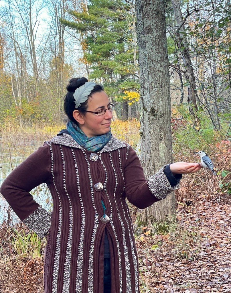 Me standing in an autumn wood with a body of water behind me. I'm holding out a hand; perched on its tip is a white-breasted nuthatch.
