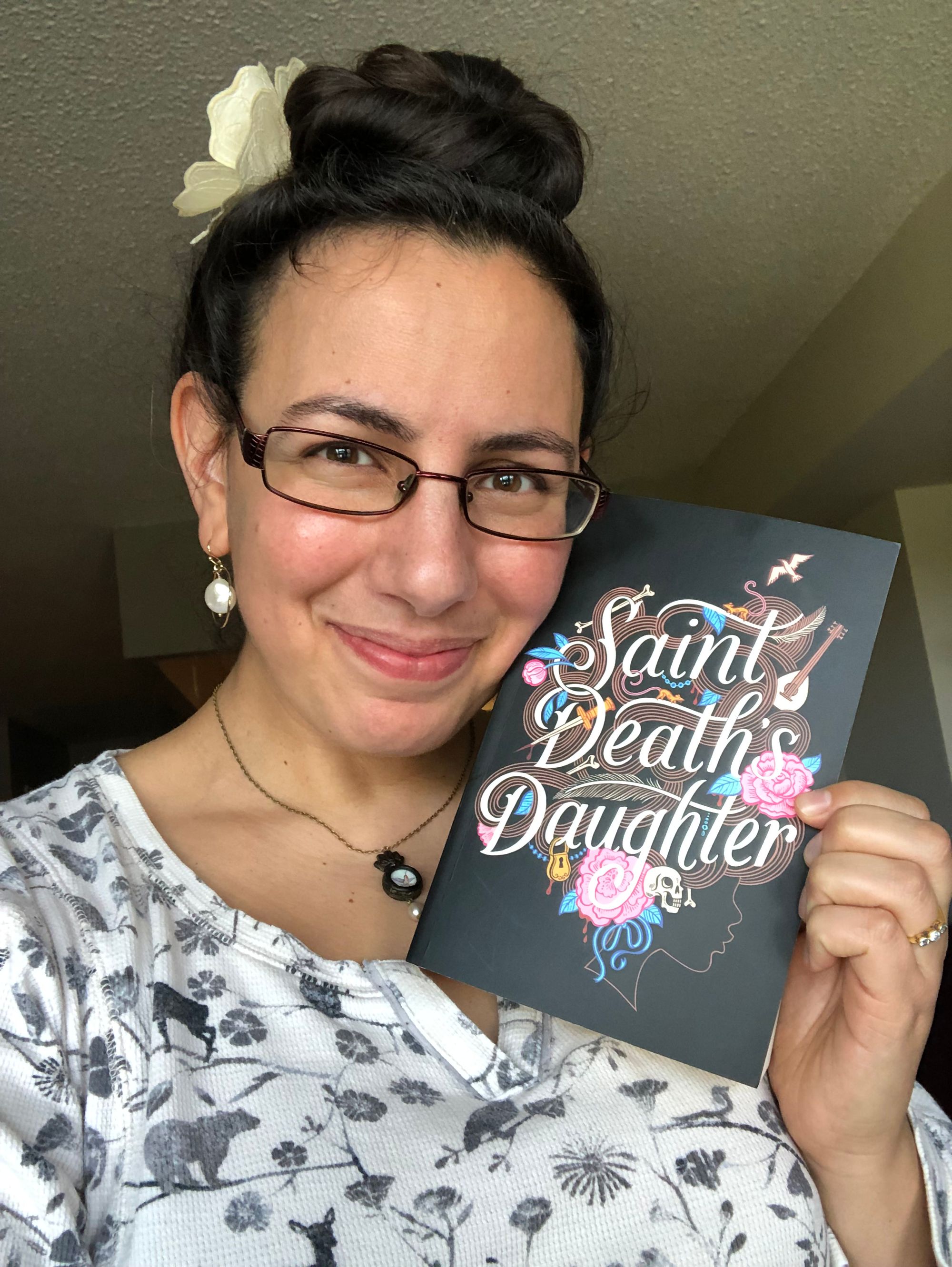 A selfie in which I tenderly rest my cheek against an advanced reading copy of C. S. E. Cooney's SAINT DEATH'S DAUGHTER while looking at the camera. Cover description: against a black background, the title words are in a cursive italic script embellished with pink roses, blue foliage, a dagger, bones, musical instruments, a padlock and a bird in flight. These design elements make up the tangled brown hair of a young woman's profile outlined in brown against the black.