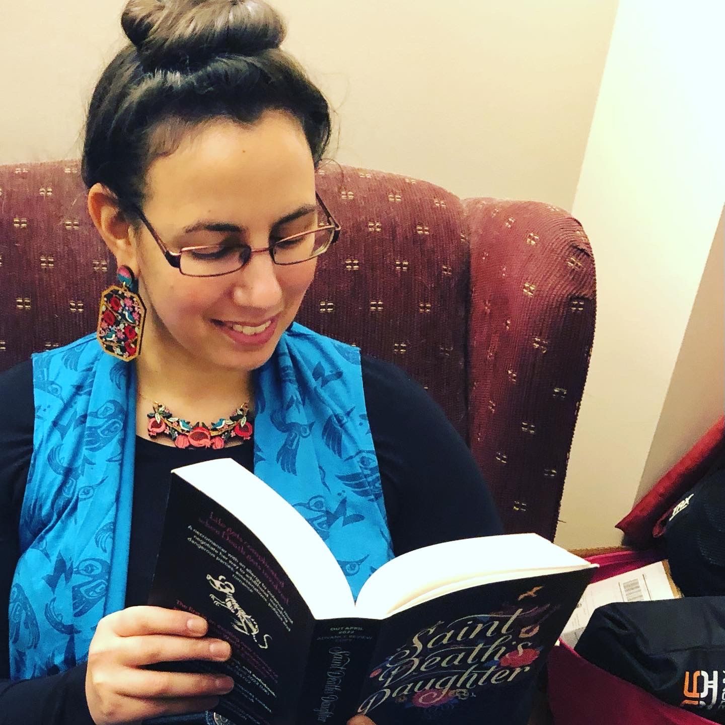 Photo of me reading Saint Death's Daughter while wearing frankly extraordinary statement earrings and necklace from Rylee and Ink, which are sculptural layers of lasercut wood handpainted to depict pomegranates, flowers, and bluebirds in bright jewel tones. 