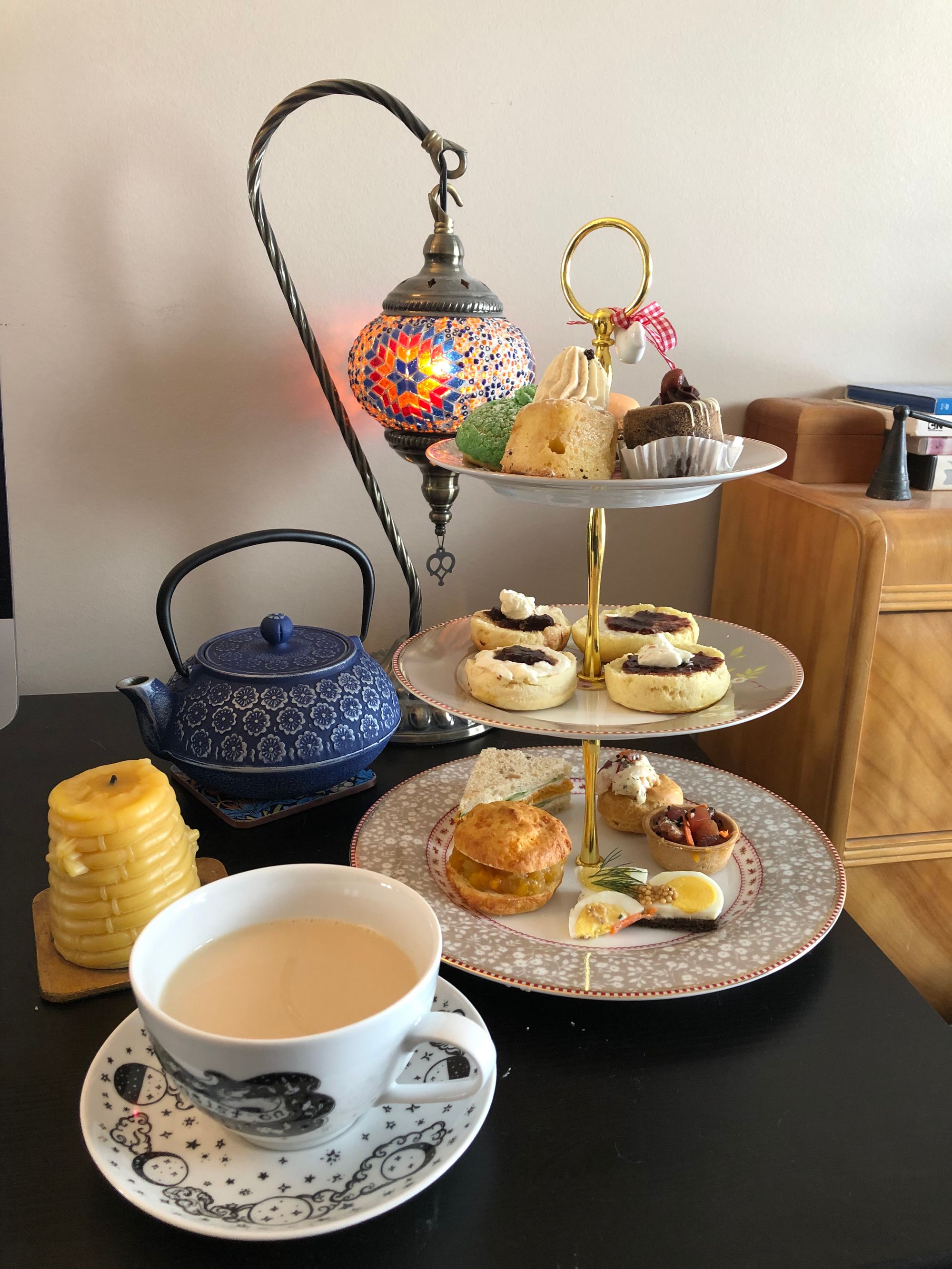 On a black desk, the following items from foreground to background clockwise: a tea cup and saucer from Cat Mallard, with a moon phase design on the baes and the words "insist on your cup of stars" around the cup; a beeswax candle in the shape of a medieval hive; a cast-iron teapot, blue with a repeating flower pattern; a Moroccan-style glass mosaic lamp, lit; a 3-tiered tray of treats, with savoury sandwiches on the bottom, halved scones with mascarpone whipped cream and strawberry jam on the middle, and an assortment of cakes at the top.