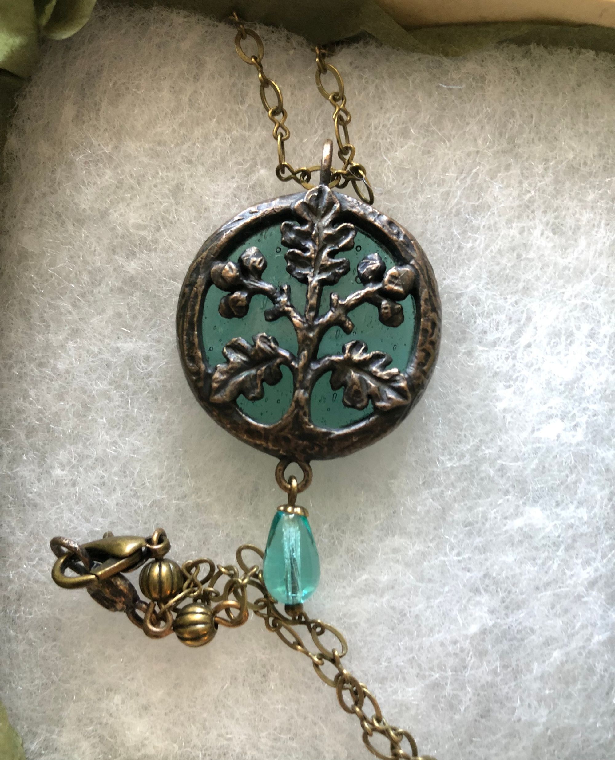 Close-up on a pendant made of antiqued metal encircling turquoise glass, resting in cotton batting. Covered in bronze patina, the circle of the pendant frames a stylized oak branch made of the same metal: three leaves point upwards and to either side, and between them grow two clusters of acorns. This figure is set over the turquoise glass. There's a turquoise glass bead dangling from the bottom of the pendant, and a bronze-coloured chain is visible above and below it as it lies flat.