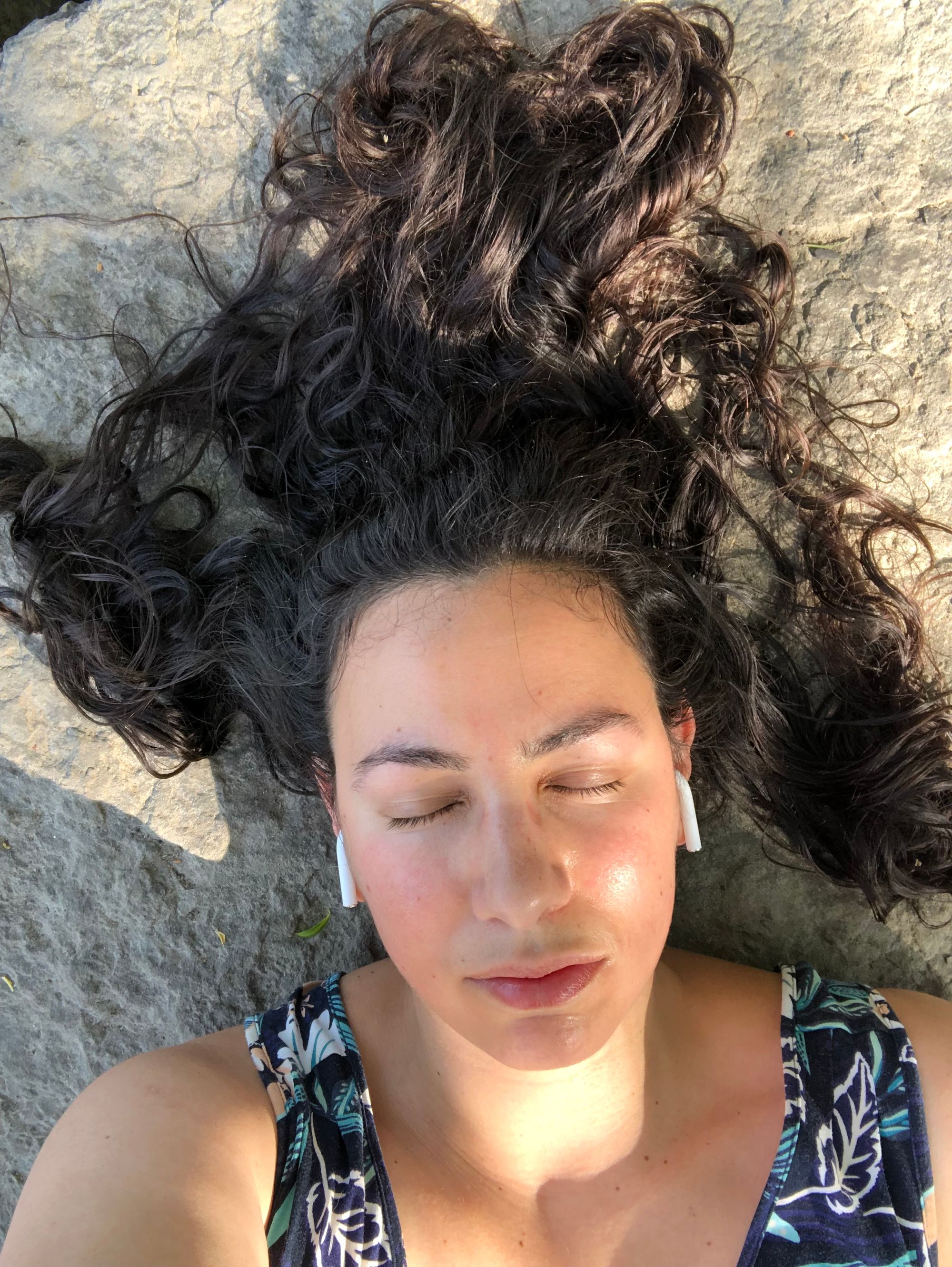 angled from above my face, a photo of me lying against grey stone with my eyes closed, my very long dark hair in wild snaking coils around my head, dappled sunlight at my throat. Wireless headphones are visible in my ears.
