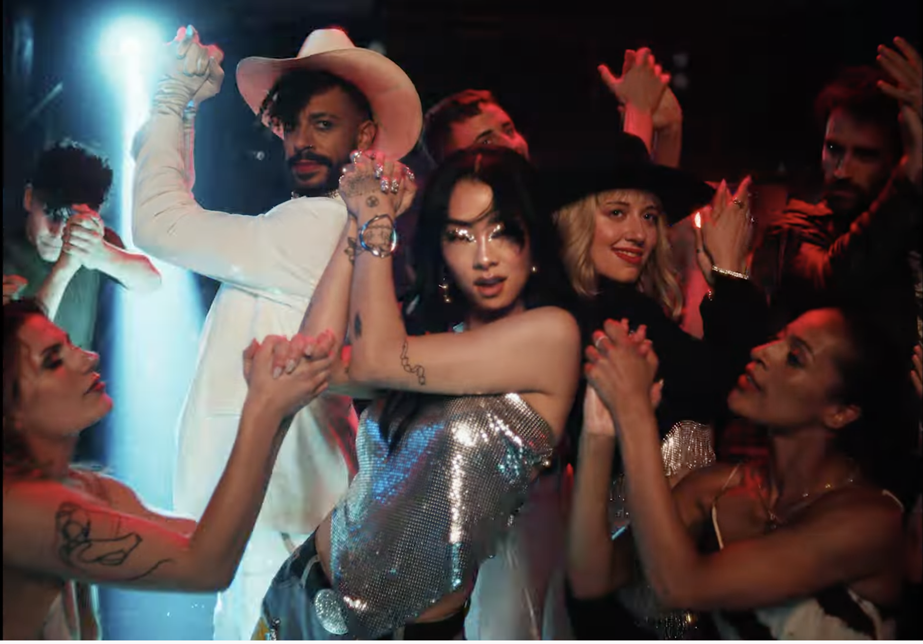 Screencap from the end of the video to "This Hell," an ensemble shot centering Rina Sawayama in a metallic strapless top with a triangular hem, her hands raised to her head at the end of a clap, wearing metallic eye makeup that echoes the top, looking fierce & joyful.