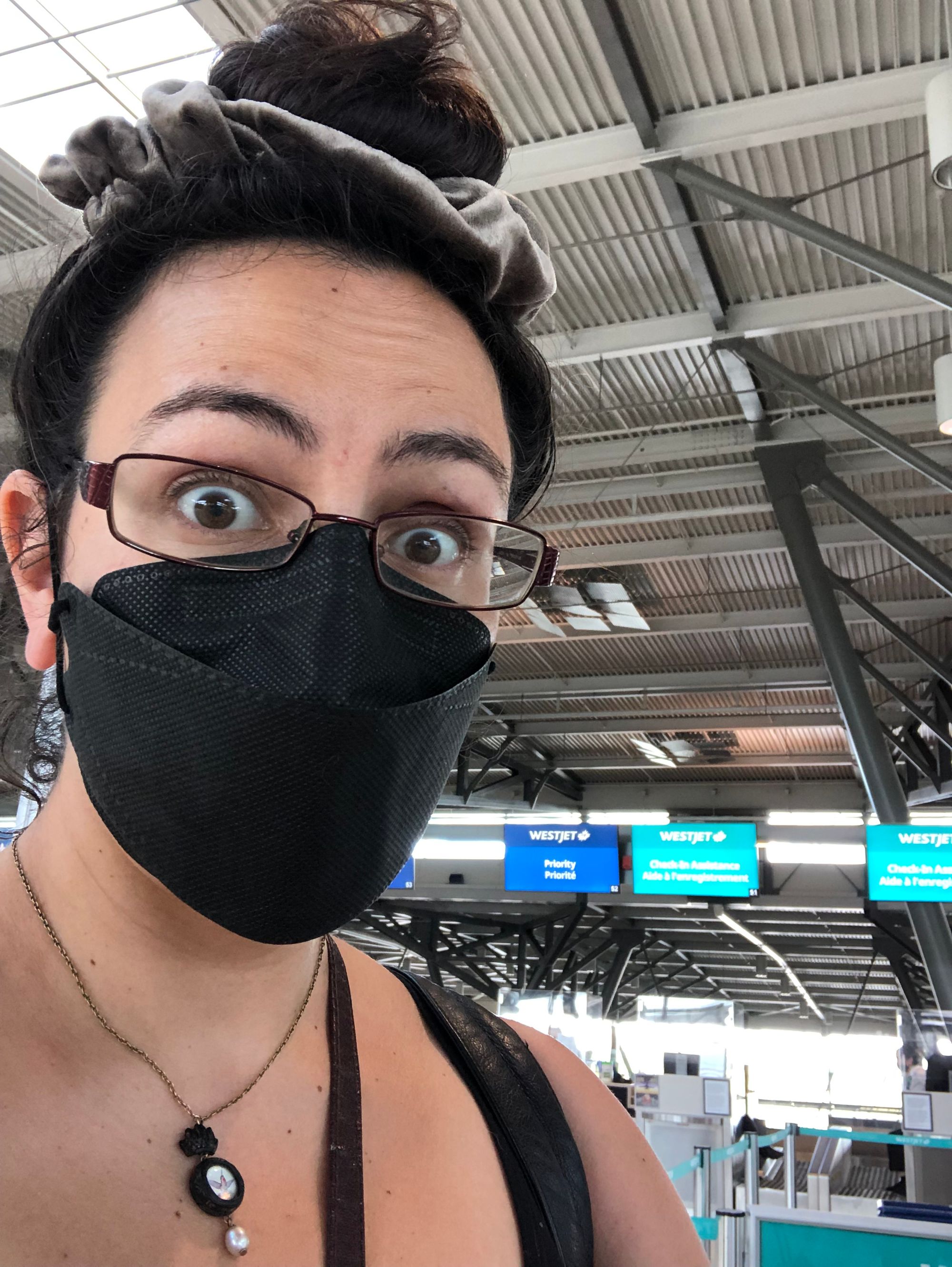 Selfie depicting a very startled-looking me wearing a black KN94 mask, a Parrish relics hummingbird pendant with a pearl drop, and my dark brown hair up in a bun surrounded by a grey velvet scrunchie. Behind me are the check-in counters for Westjet at the Ottawa International Airport.