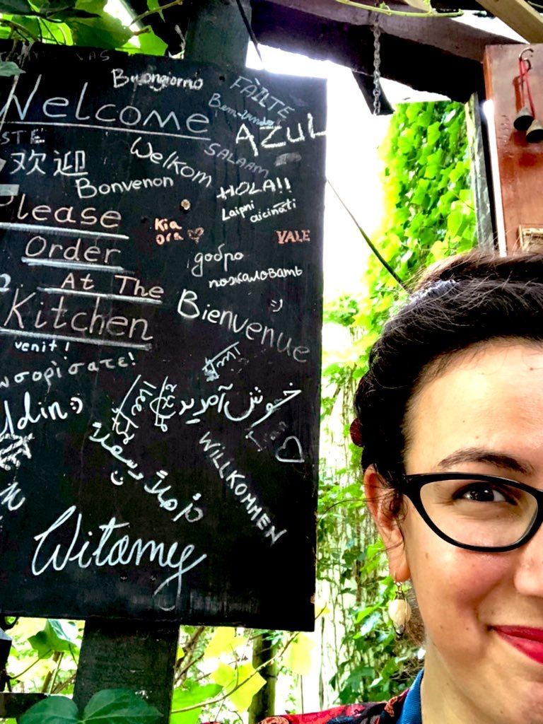 A selfie in which only half my face is visible at bottom right, most of the frame given over to a blackboard surrounded by greenery on Tchai Ovna's outdoor patio, on which "welcome" is written in several different languages including Arabic, Greek, French, Maori, Spanish, Italian and German, surrounding the English words "Please Order at the Kitchen."