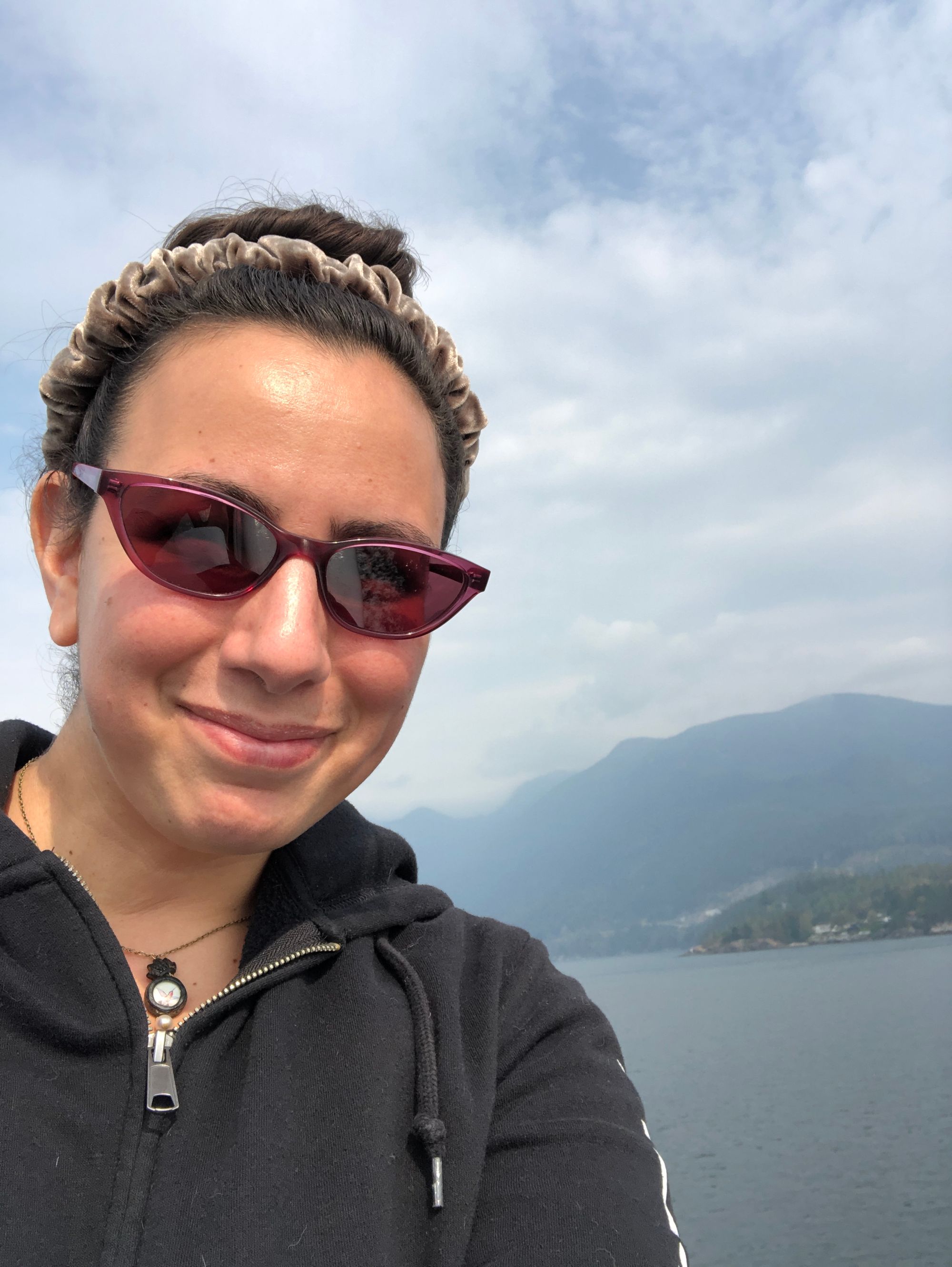 Selfie from a ferry, in which I'm wearing sunglasses and a scrunched velvet headband, with water and mountains behind me. 