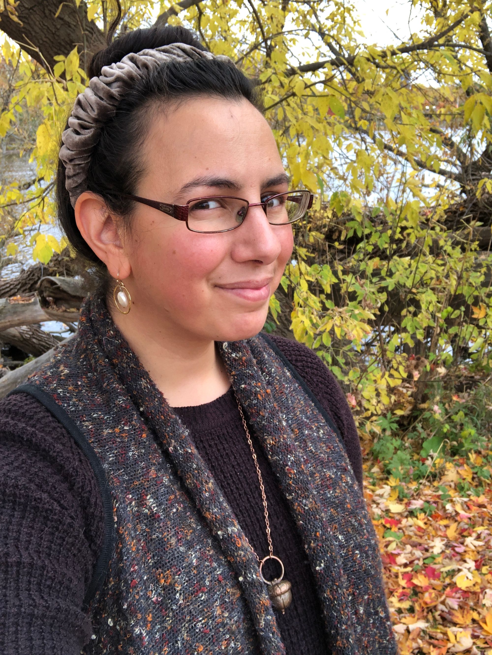 Selfie depicting me in a three quarter turn to camera, smiling with mouth closed, wearing rectangular glasses with a metal frame and my dark hair in a high bun. Behind me is a colourful mess of autumn leaf-fall on the ground and a yellow-green drapery of leaves on trees, beyond which is a river. My outfit's autumnal in texture and colour: a thick dark brown rib-knit sweater with a long sleeveless cardigan atop it, flecked with colours that pick up on the leaf-fall, and accessorized with the following jewelryL a long copper-coloured necklace that ends in a bronzed acorn; flat pearl earrings floating in a ring of gold; and a scrunched grey-brown velvet headband.