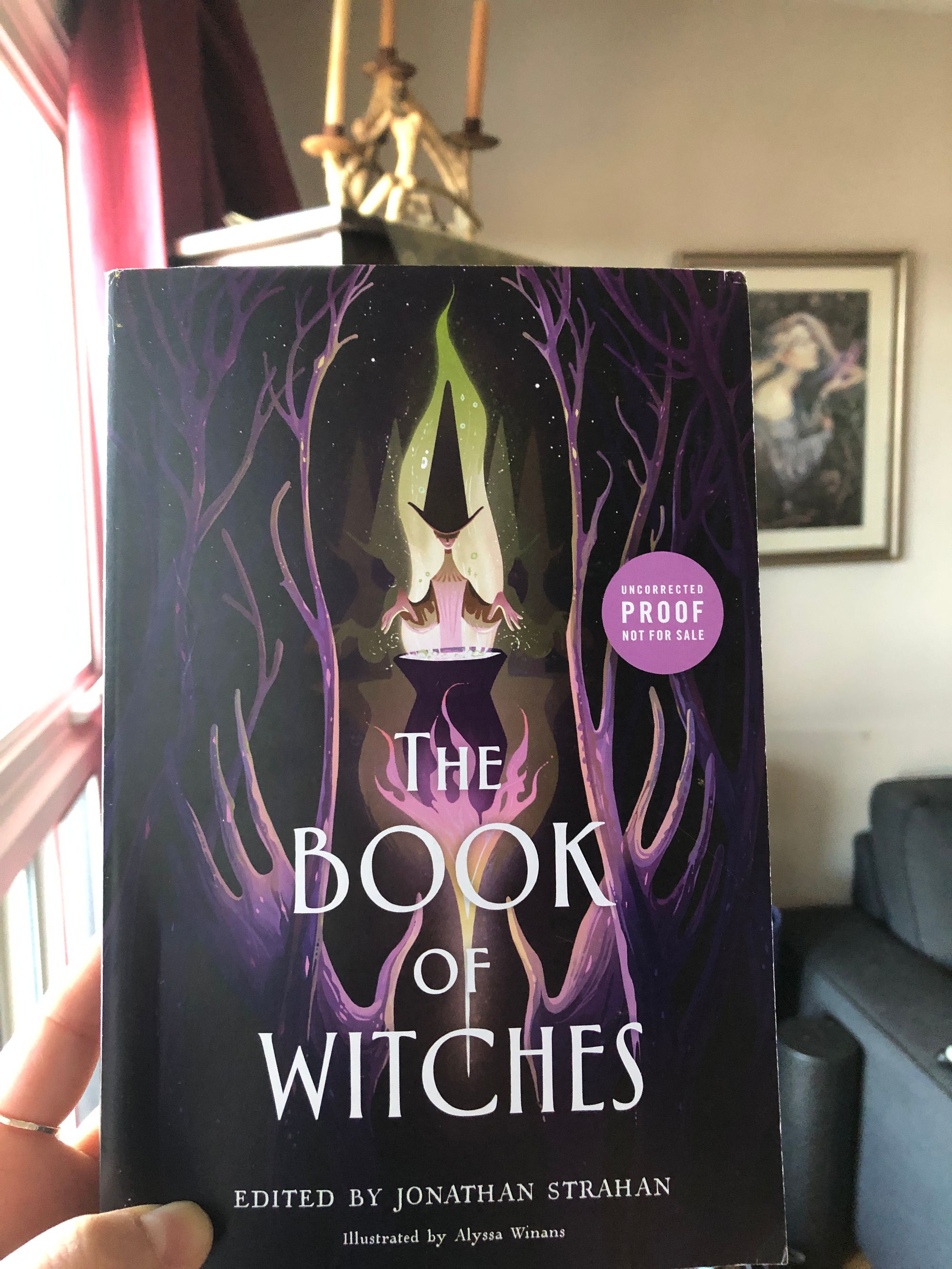 Close up on the cover of THE BOOK OF WITCHES, edited by Jonathan Strahan, held up in my hand. The illustration by Alyssa Winans depicts a stylized witch presiding over a bubbling cauldron in a clearing in the woods, all drawn in a slender, spooky cartoonish style in purples and acidic greens; the woods in the foreground look like slender-fingered hands reaching towards the central figure, whose own hands hover over the cauldron, from which a wispy green glow emanates, framing her.