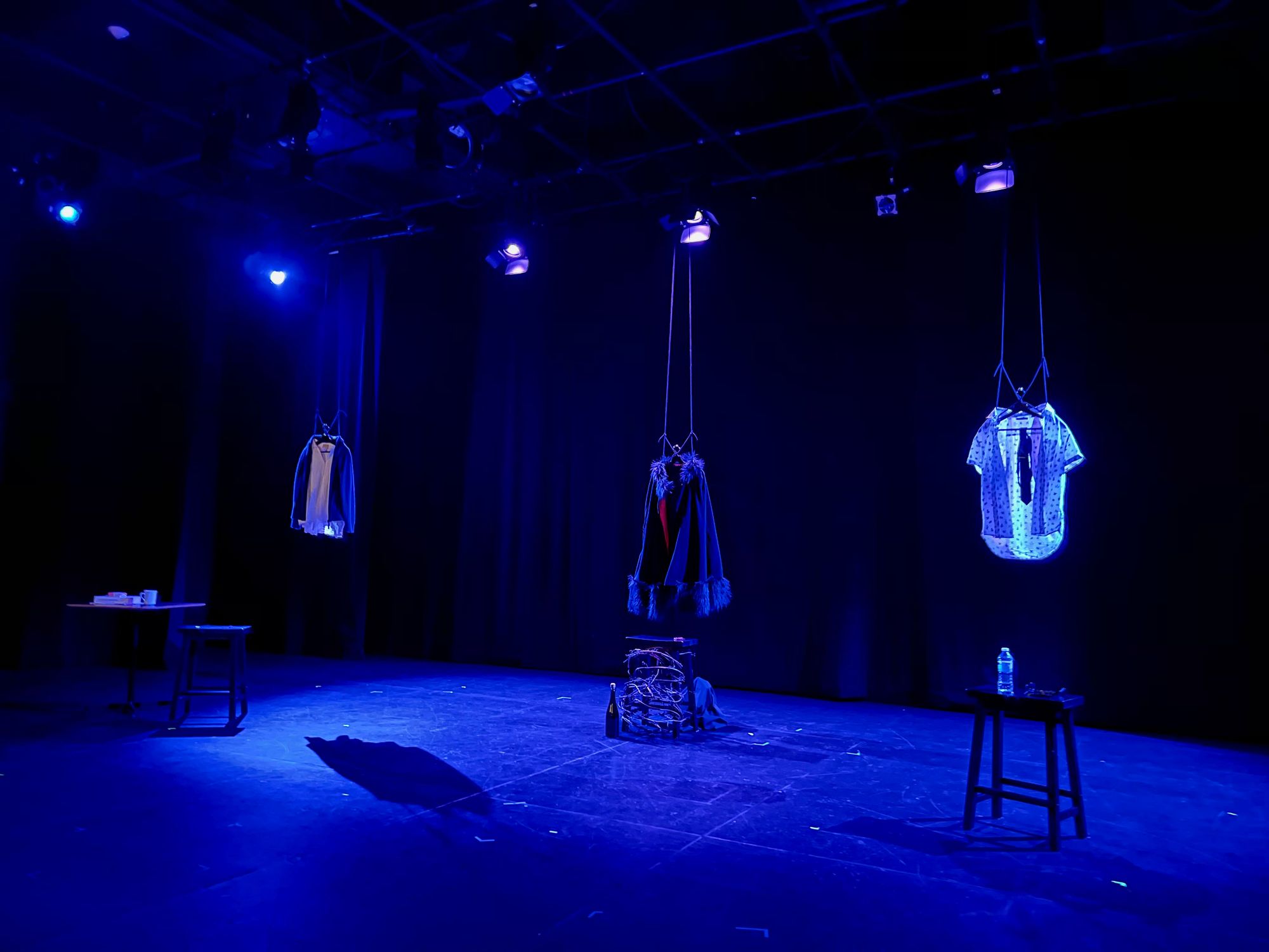 The empty stage for DRESSED AS PEOPLE: AN UNCANNY TRIPTYCH. The stage is lit in cool blues and set with three minimalist stations for each monologue, each marked out by an outfit hung from the ceiling. To the far left is a modest shirt and cardigan next to a stool; in the middle is a fur-lined cloak atop a stool wrapped in brambles next to a bottle of wine; to the right is a snazzy short-sleeved button-up shirt with tie, over a stool set with glasses and a water bottle.
