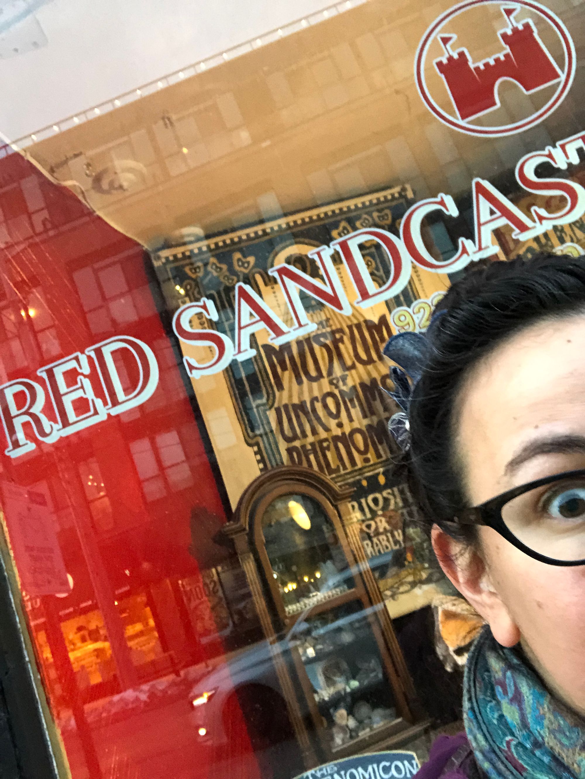 An angled photo capturing one eye and part of my face standing in front of the Red Sandcastle Theatre’s window—I’m trying to fit the whole logo and my face but simply cannot.