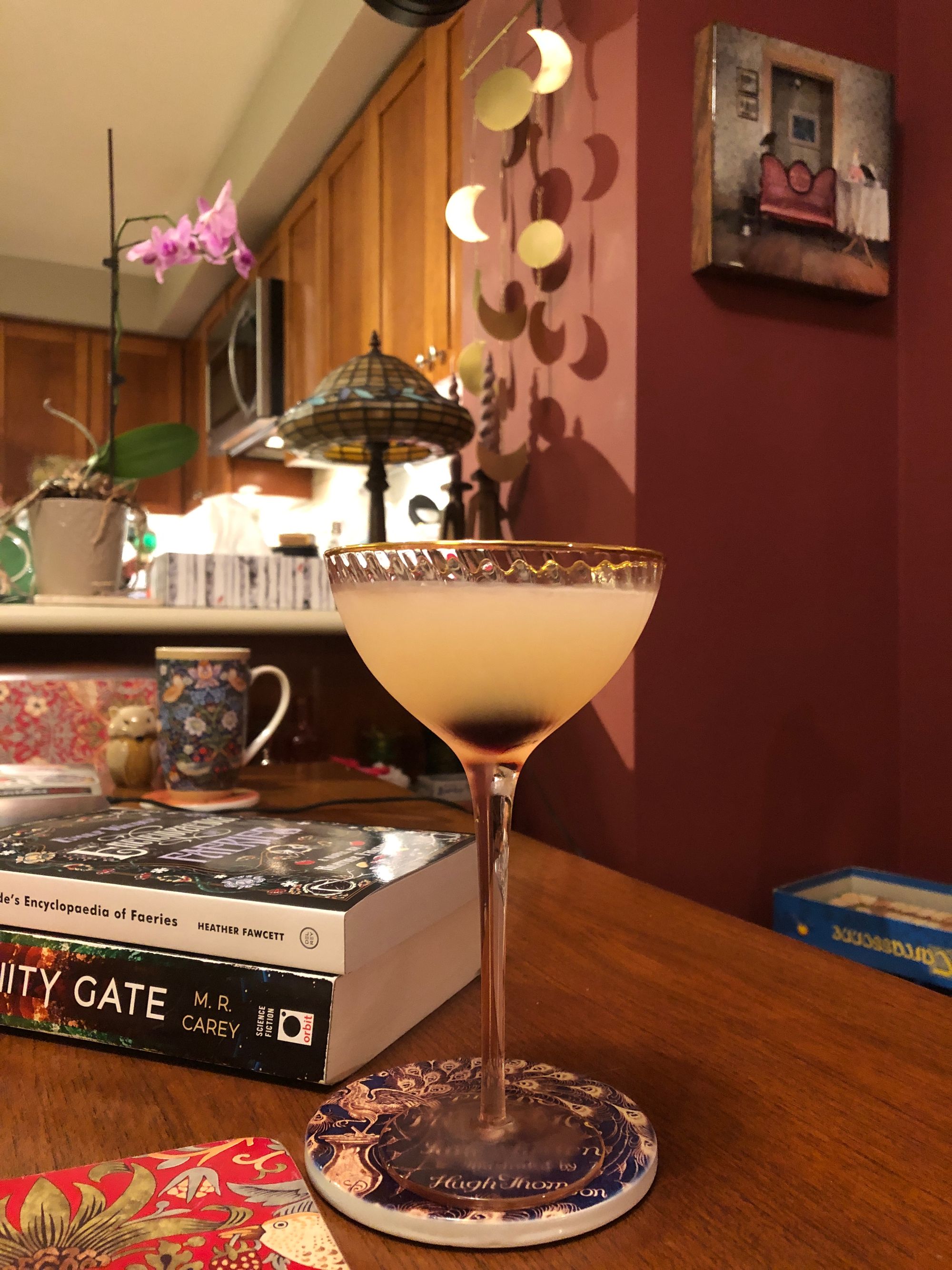 Foregrounded is an ornate coupe full of a pale cocktail with a dark cherry blooming up from the stem; the glass is set on a ceramic coaster with classical peacock imagery, next to a place mat covered in William Morris' "Strawberry Thief" print. In the background is a small stack of books, a tea cup with another Morris print, a ceramic salt shaker in the shape of a fox, a pink orchid, a stained-glass lamp, and a cascade of ornamental brass moons in various phases dangling on brass chain from a replica of a Victorian train station clock. 