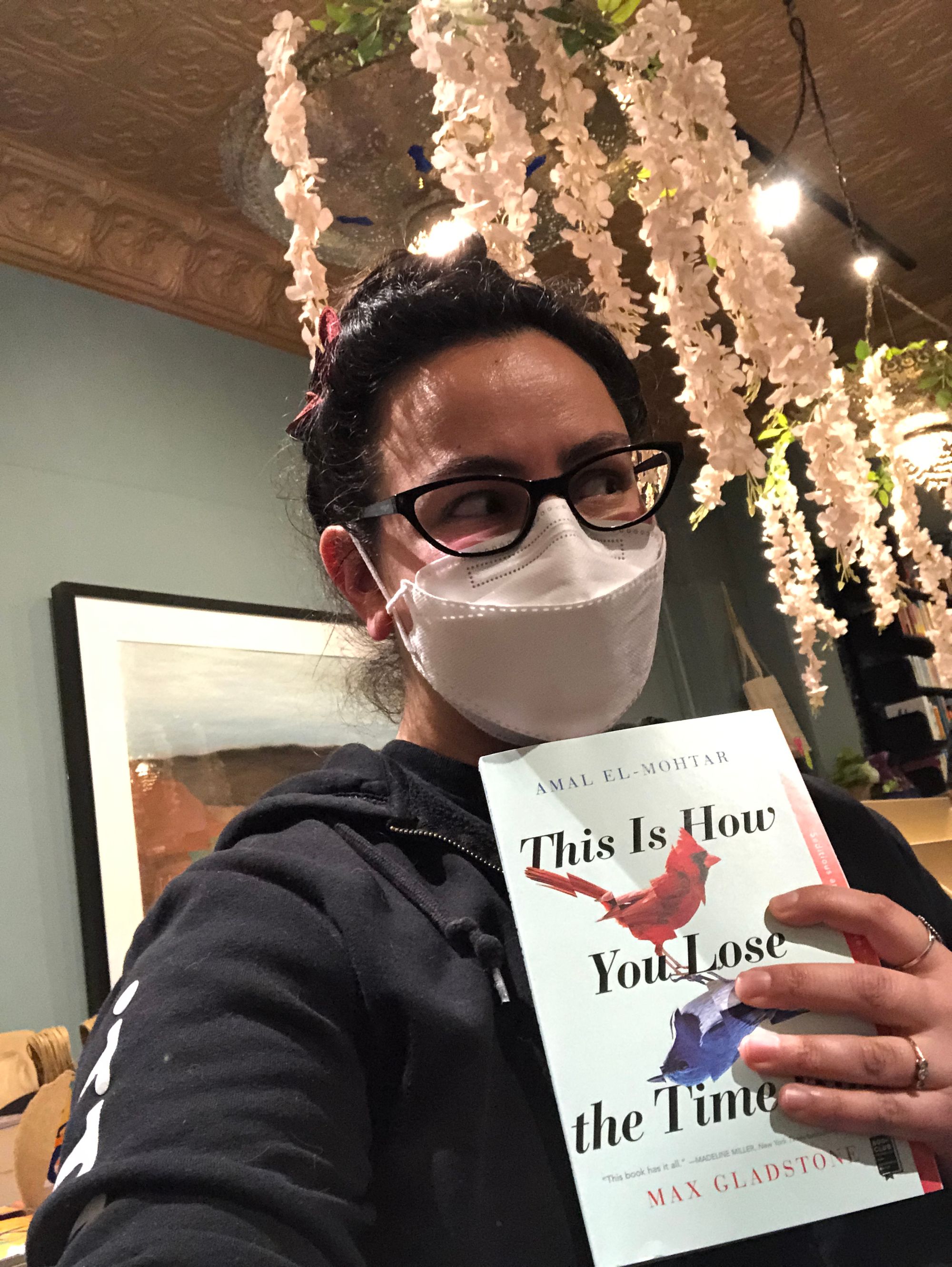 Selfie in which I'm holding up a copy of This Is How You Lose the Time War while dressed in a black hoodie and white mask, and looking slyly away from the camera like I'm doing something sneaky. Behind me are artifical drapes of wisteria dangling from the book store's light fixtures.