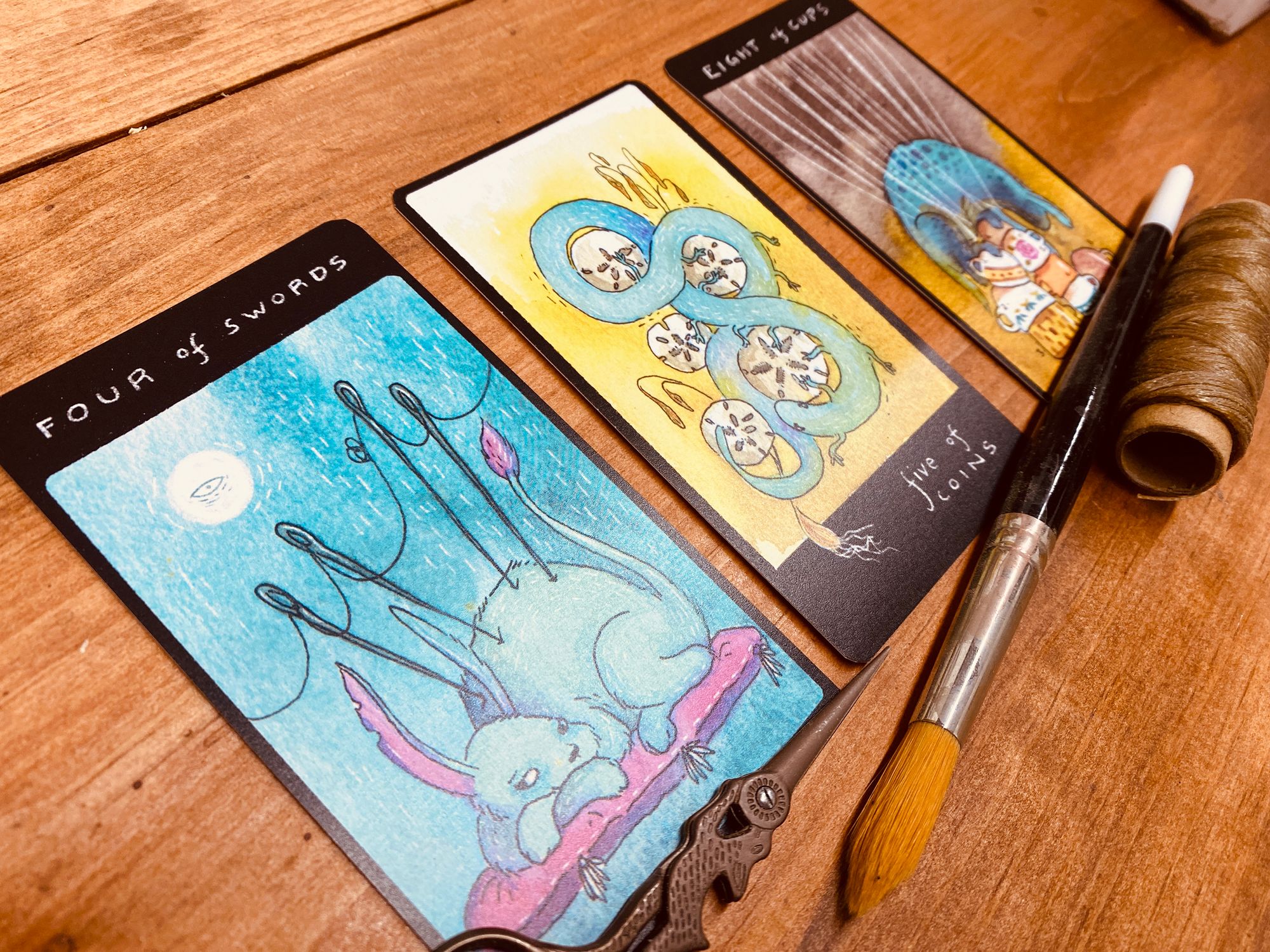 A diagonally angled shot of three tarot cards lined up over a pair of sewing scissors, a paintbrush, and a skein of brown thread. The cards are the Four of Swords (a fluffy beast with rabbit-like ears and a lion's tail reclining on a cushion while four threaded needles are poked upright into the central mass of its body), the Five of Coins (a serpentine, salamander-like creature encircles five sand dollars) and the Eight of Cups (a little blurred out, but a spotted reptile-like creature perches atop a stack of colourful tea cups.)