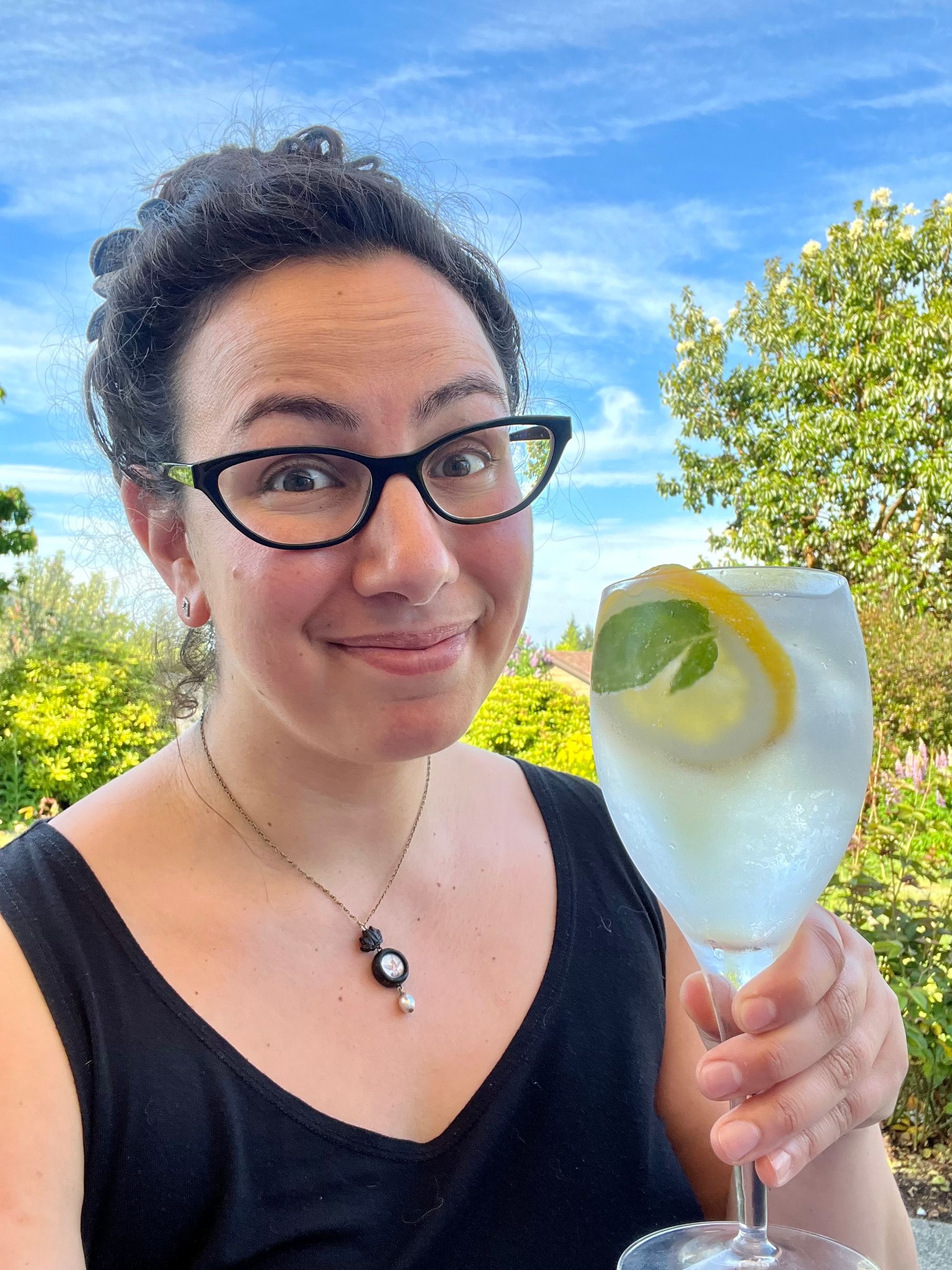 Selfie in which I look bemused but happy, in a brightly lit garden against a blue sky with white streaky clouds, holding up a wine glass full of gin and tonic with lemon and mint. 