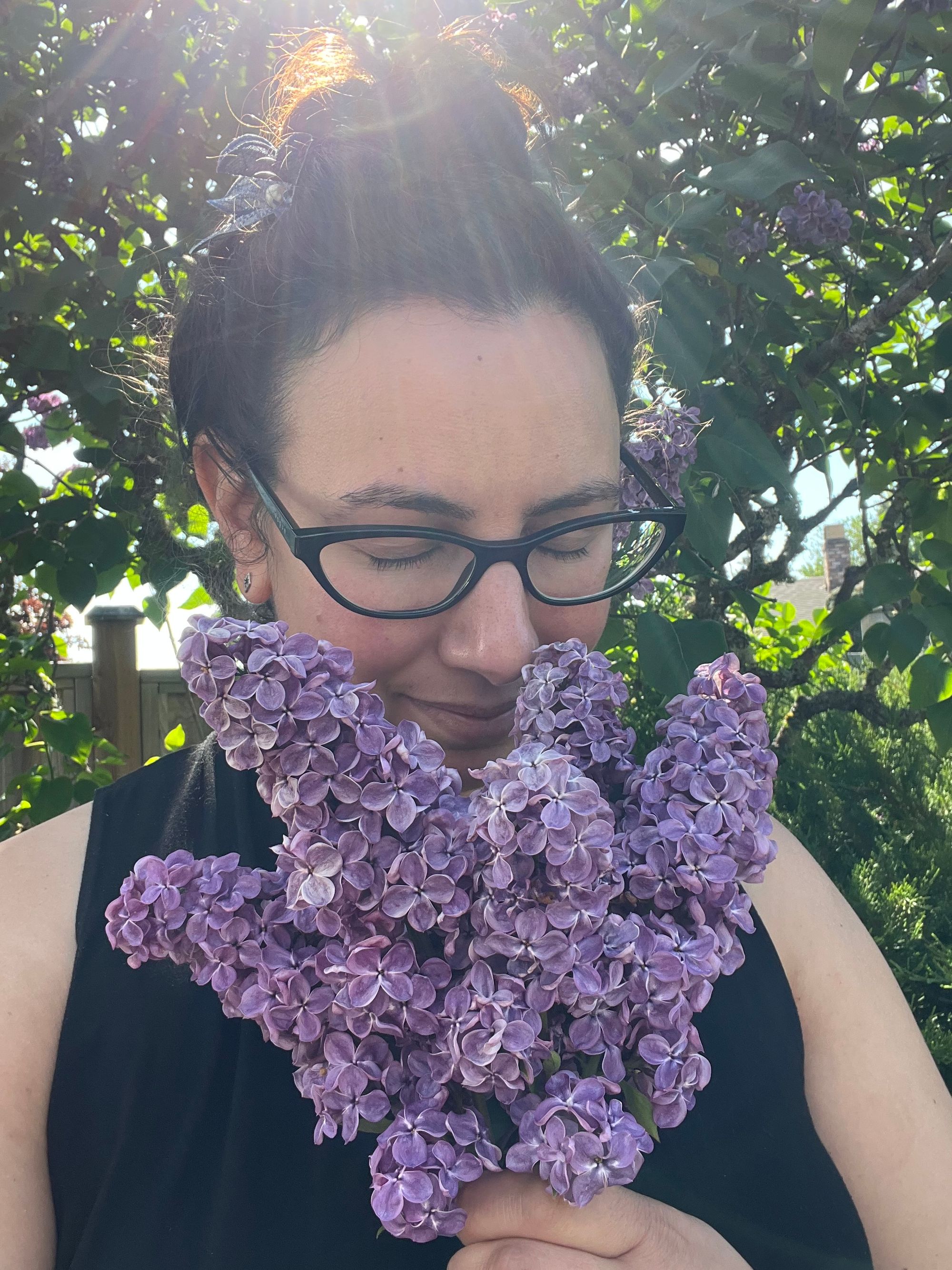 Selfie of me with eyes closed, holding up a bunch of lilacs to my nose while showered in sunlight.
