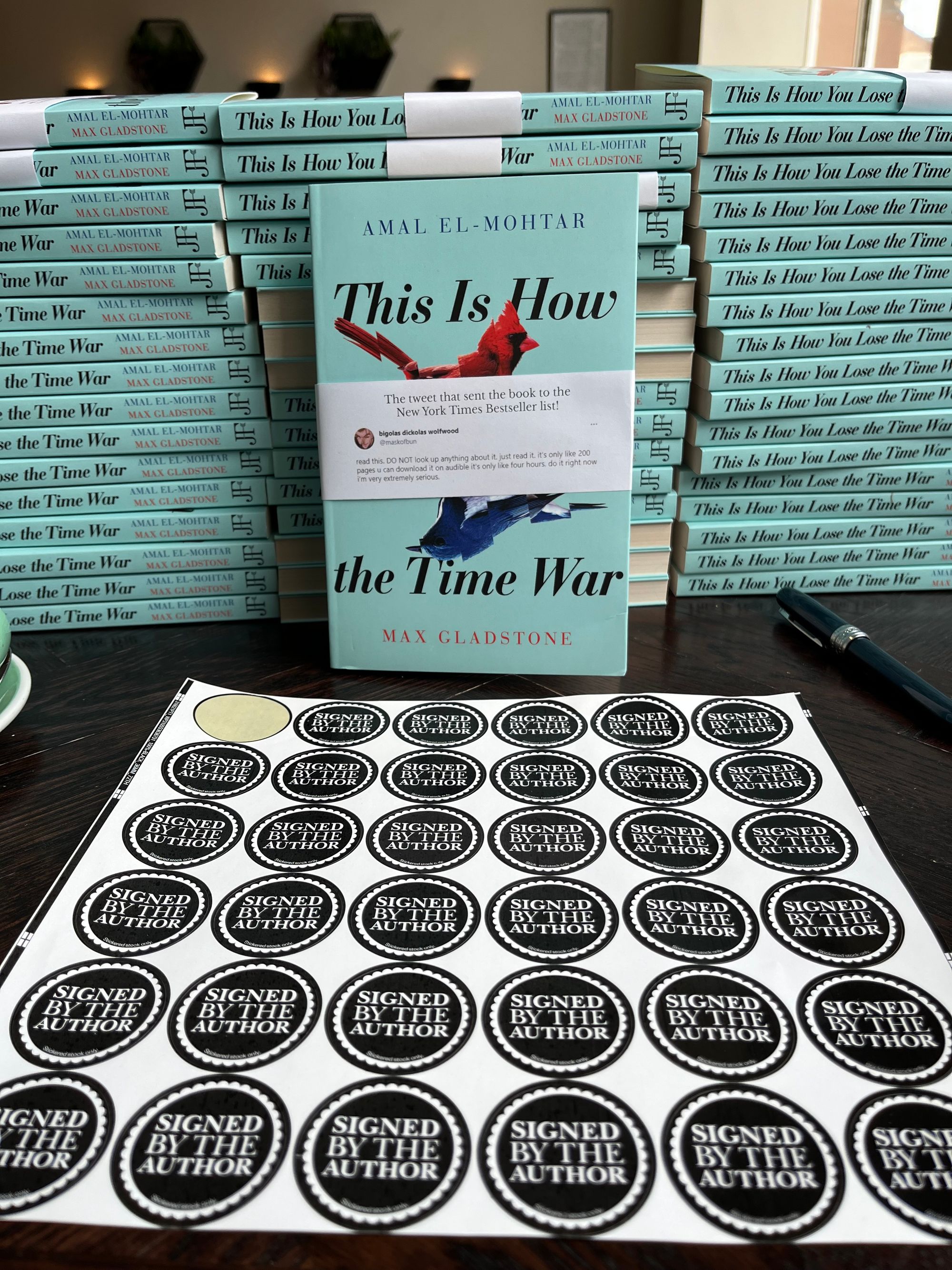 Roughly fifty paperback copies of This Is How You Lose the Time War arranged in three equal stacks on a table, with one copy faced out in front of them, displaying a printed sleeve of Bigolas Dickolas Wolfwood’s infamous tweet recommending the book. In the foreground is a sheet of Signed by the Author stickers to be put on the books once I’ve signed them.
