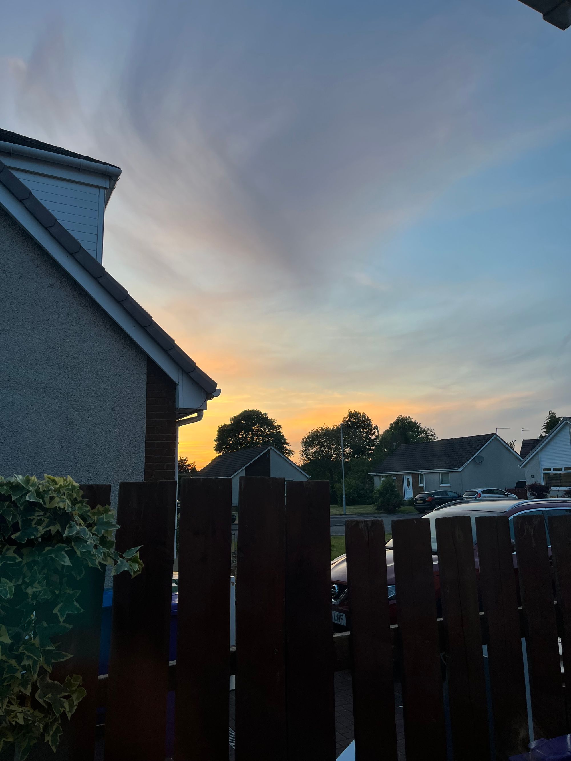 An evening sky glimpsed over a wooden fence and rooftops of detached houses, still full of the bright gold of a sunset's afterglow.