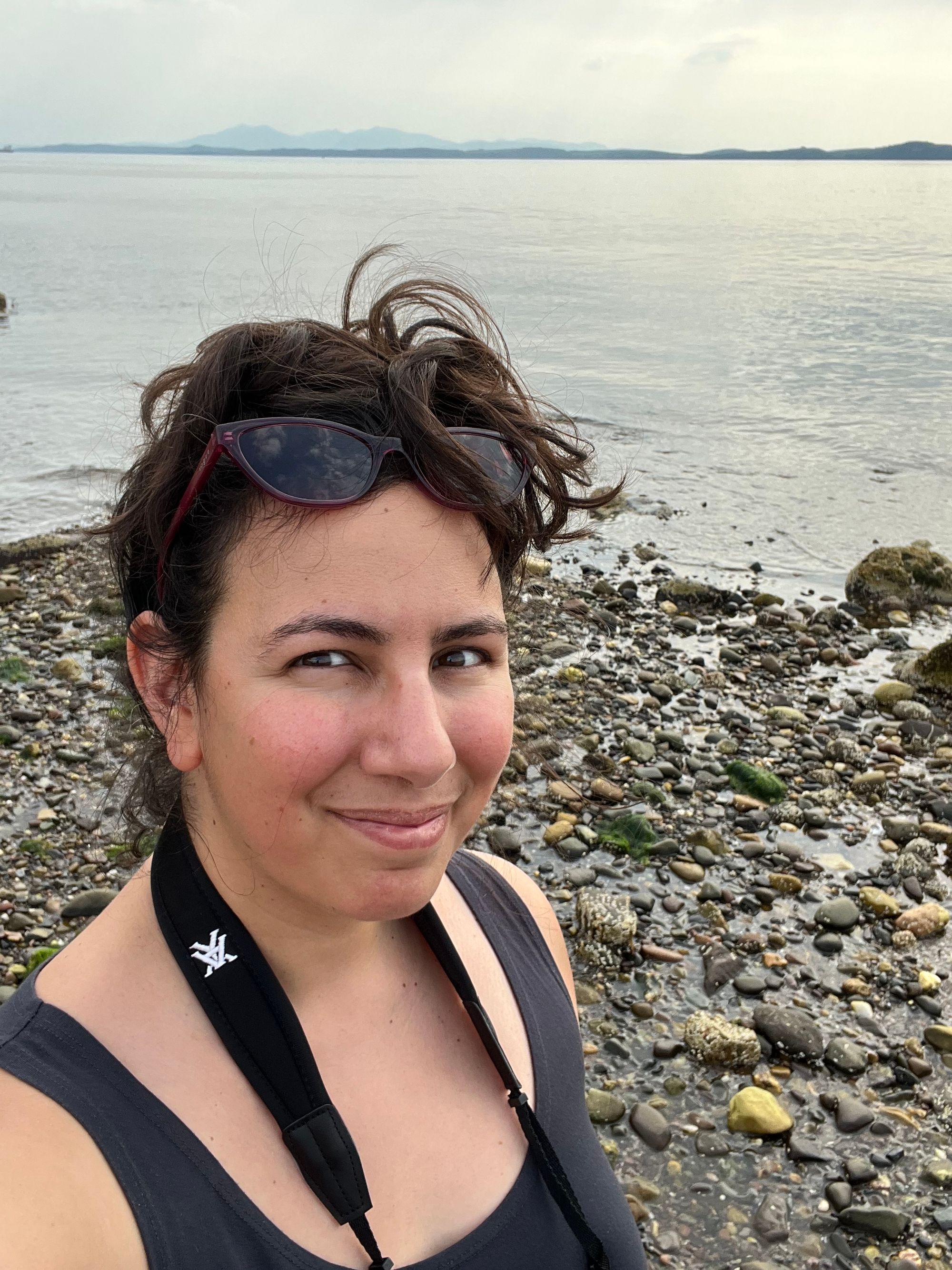 Selfie in which I'm wearing my sunglasses on top of my head amid a very messy top knot of dark hair, and binocular straps over a dark grey tank top, against a backdrop of grey sea and distant mountains silhouetted in blue on the horizon.