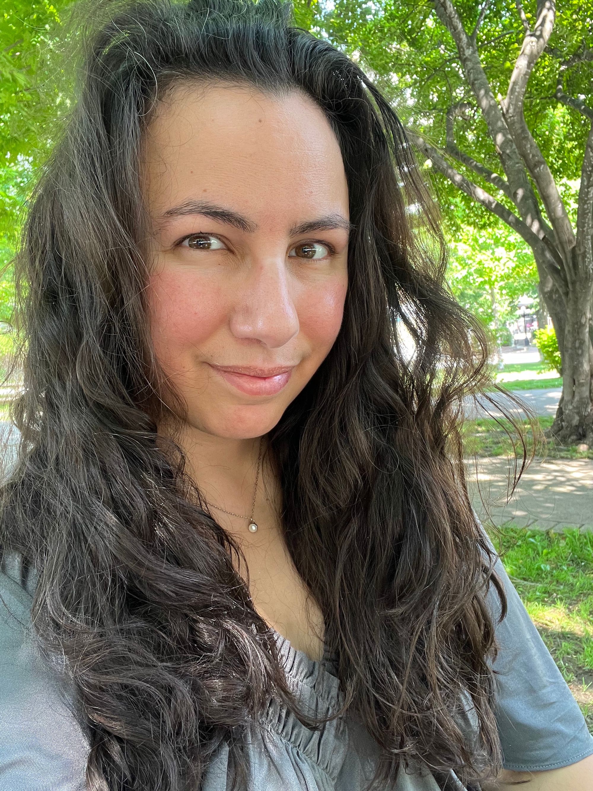 Softly lit park selfie in which I’m wearing my long dark hair loose over a grey silk top with a plunging pleated neckline and loose, fluttering sleeves.