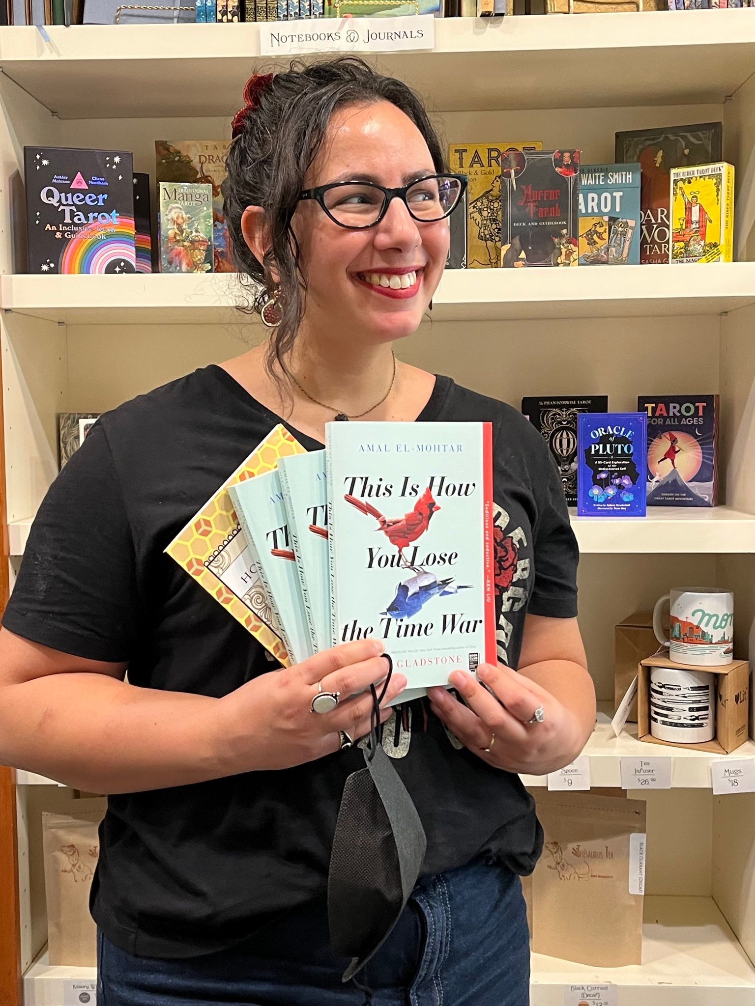 Photo of me from the waist up wearing a Hadestown t-shirt and looking off to the side, delighted and smiling, holding up three copies of This Is How You Lose the Time War and one copy of The Honey Month, fanned out together. Behind me is a shelf full of Tarot decks, tea mugs and tea. 