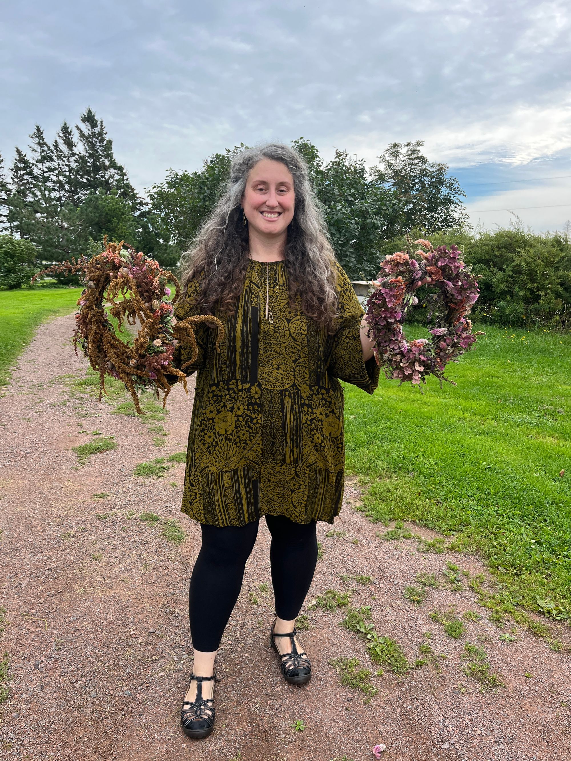 A smiling Caitlyn, wearing a green-and-black tunic in a foliate pattern over black leggings, holds up two flower crowns, one mostly amaranth in warm tones and the other mostly celosia in cooler tones.