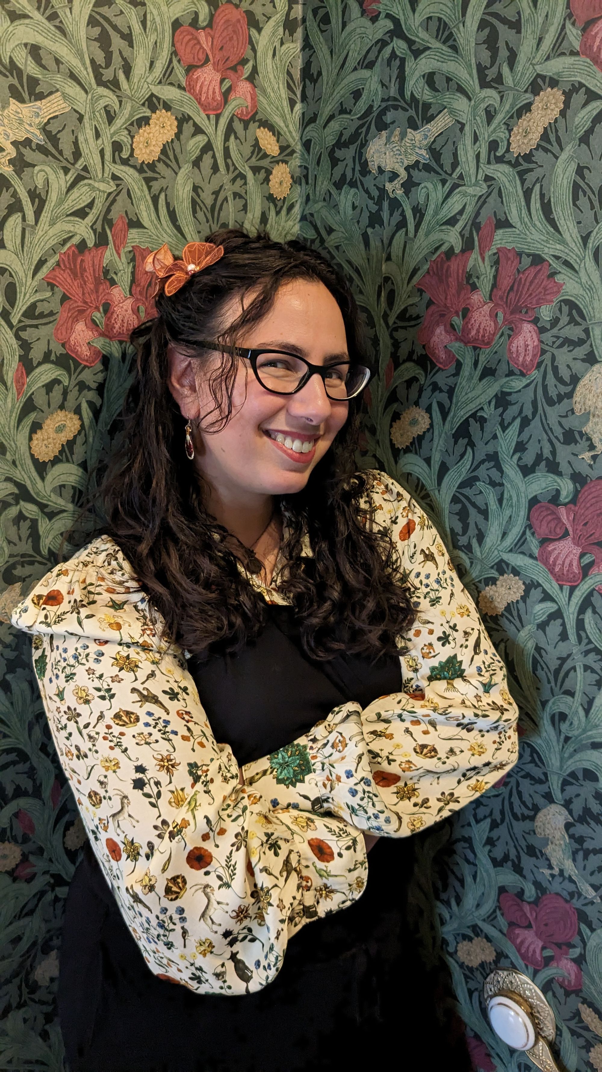 Photo of me in a beautifully wallpapered corner. The wallpaper is foliate, lush greens against a dark green background with pops of purple irises and pale silhouettes of birds. I'm wearing my long dark hair dark in curling waves, with hand-painted pomegranate earrings by Luna Sangre, a red-brown silk hair flower by Betsie Withey, and a silk blouse with billowing sleeves in a medieval manuscript print, by Samantha Pleet, under a black jumpsuit. My arms are crossed and I'm smiling. 