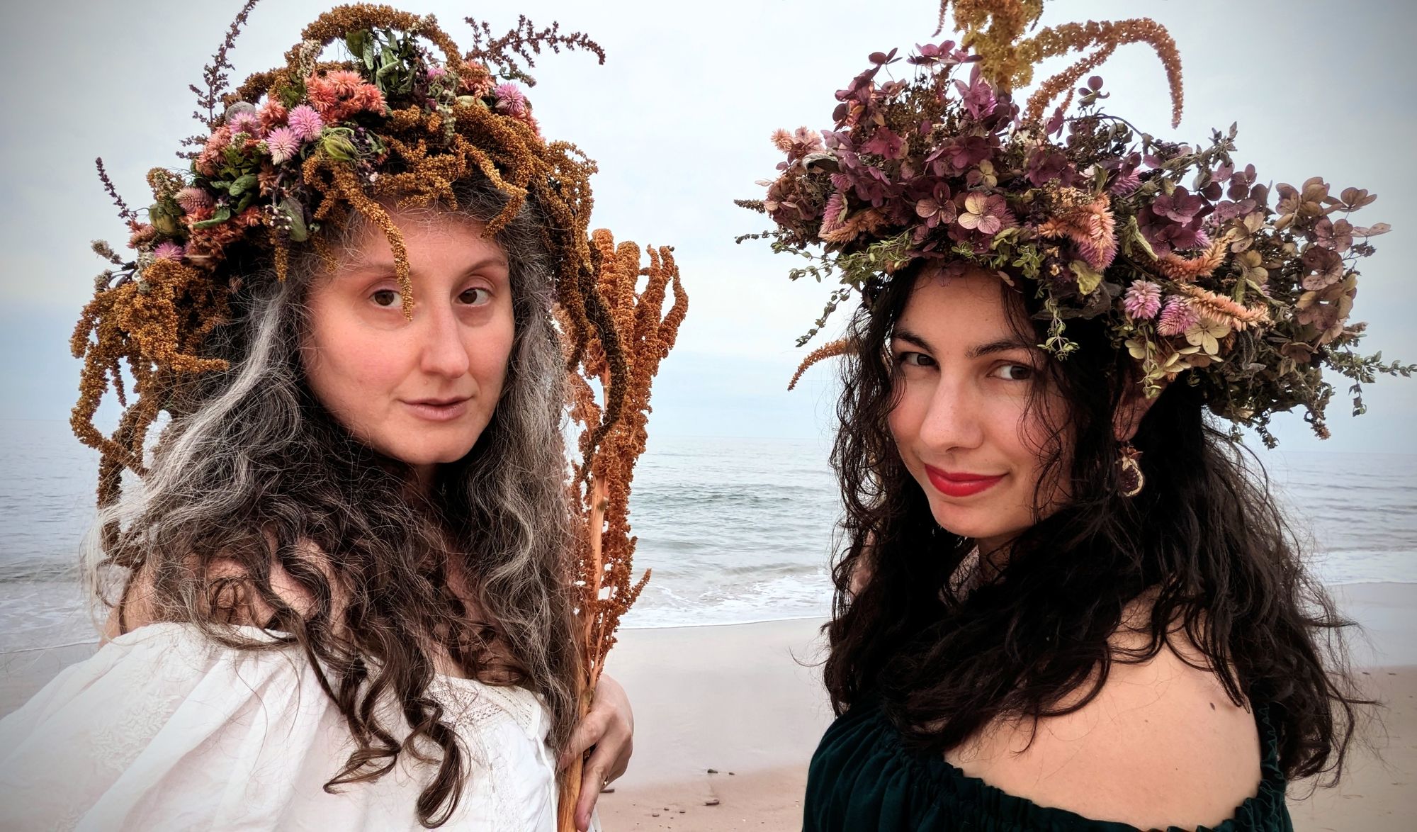 A dramatic beach photo in which two women stand across from each other wearing magnificently elaborate flower crowns and holding tall stalks of amaranth with the ocean behind them, looking over their shoulders towards the camera.  On the left is Caitlyn Paxson, a white woman with sorcerous streaks of white and silver in her long curling brown hair, wearing a flowing white shirt and a crown featuring long tendrils of amaranth; on the right is Amal El-Mohtar, a middle-eastern woman with very dark wavy hair, wearing a crown featuring celosia and hydrangea. Both women are smiling slightly and look amazing.