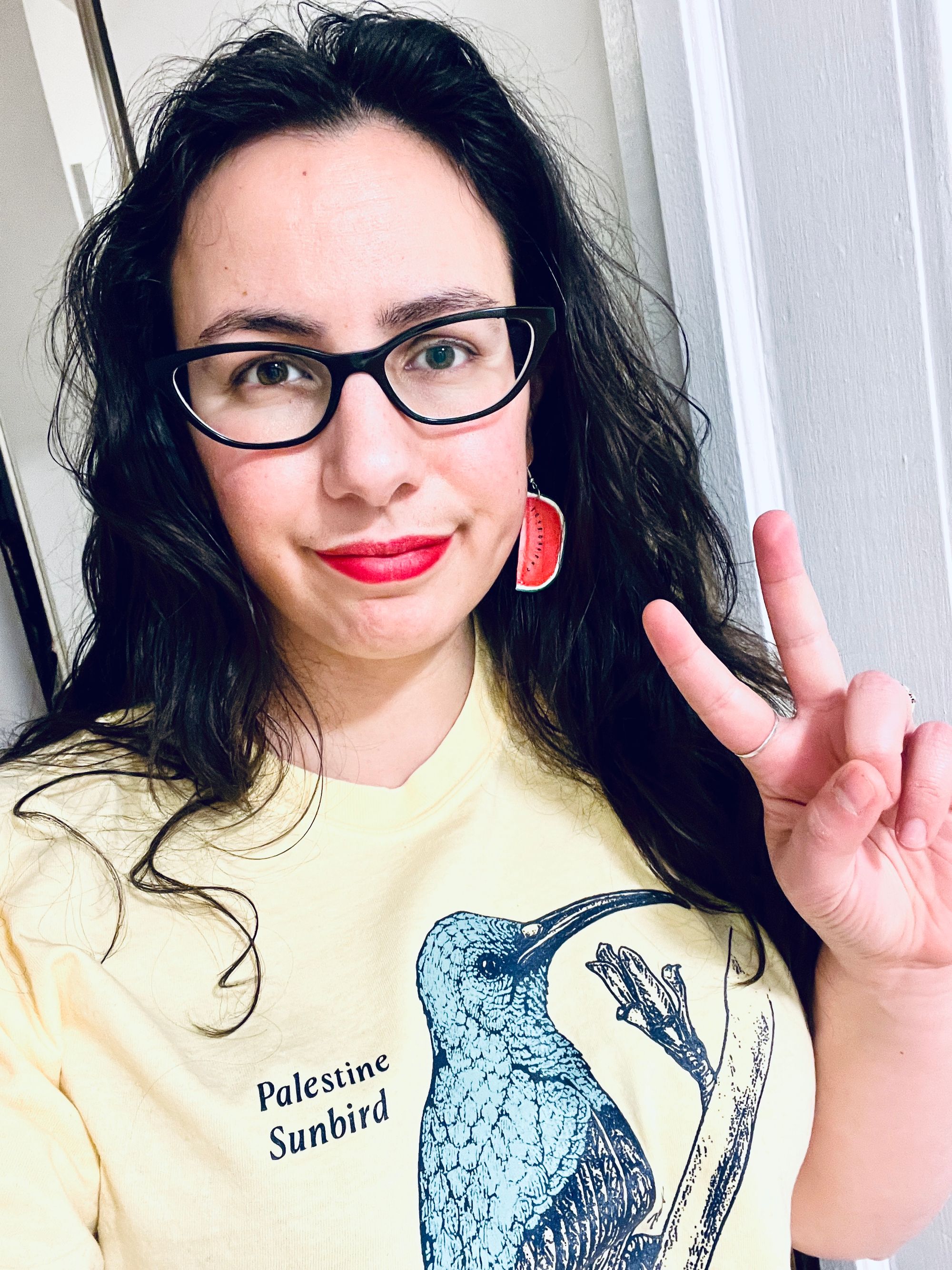 Selfie in which my dark wavy hair is down around my shoulders and spilling over a pale yellow t-shirt depicting a Palestine Sunbird. I'm wearing watermelon-slice earrings and raising two fingers in a peace sign.