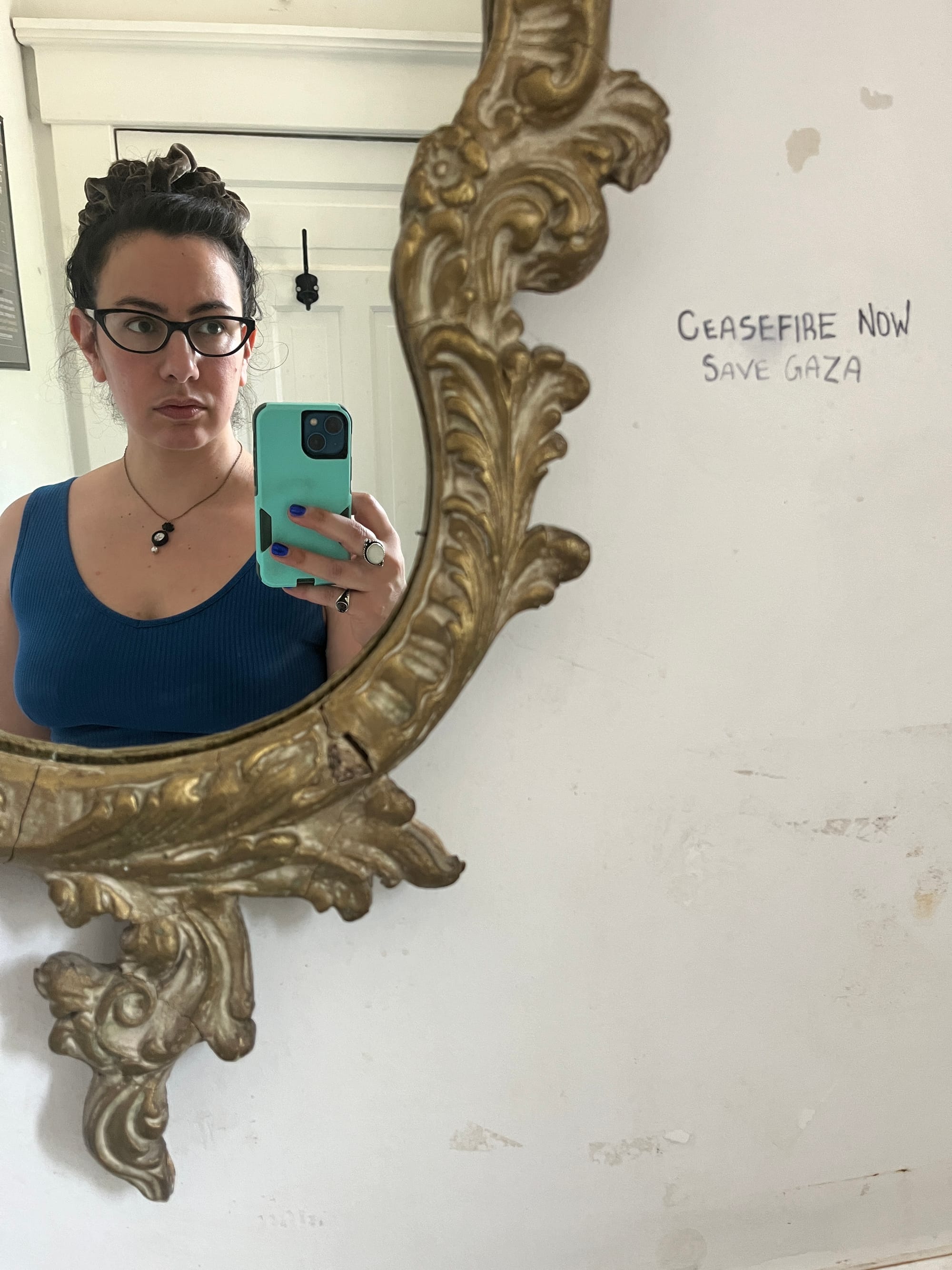 Mirror-selfie taken from the bust up in a partially visible gloriously ornate gilt mirror frame, next to which someone has scribbled “Ceasefire Now, Save Gaza”