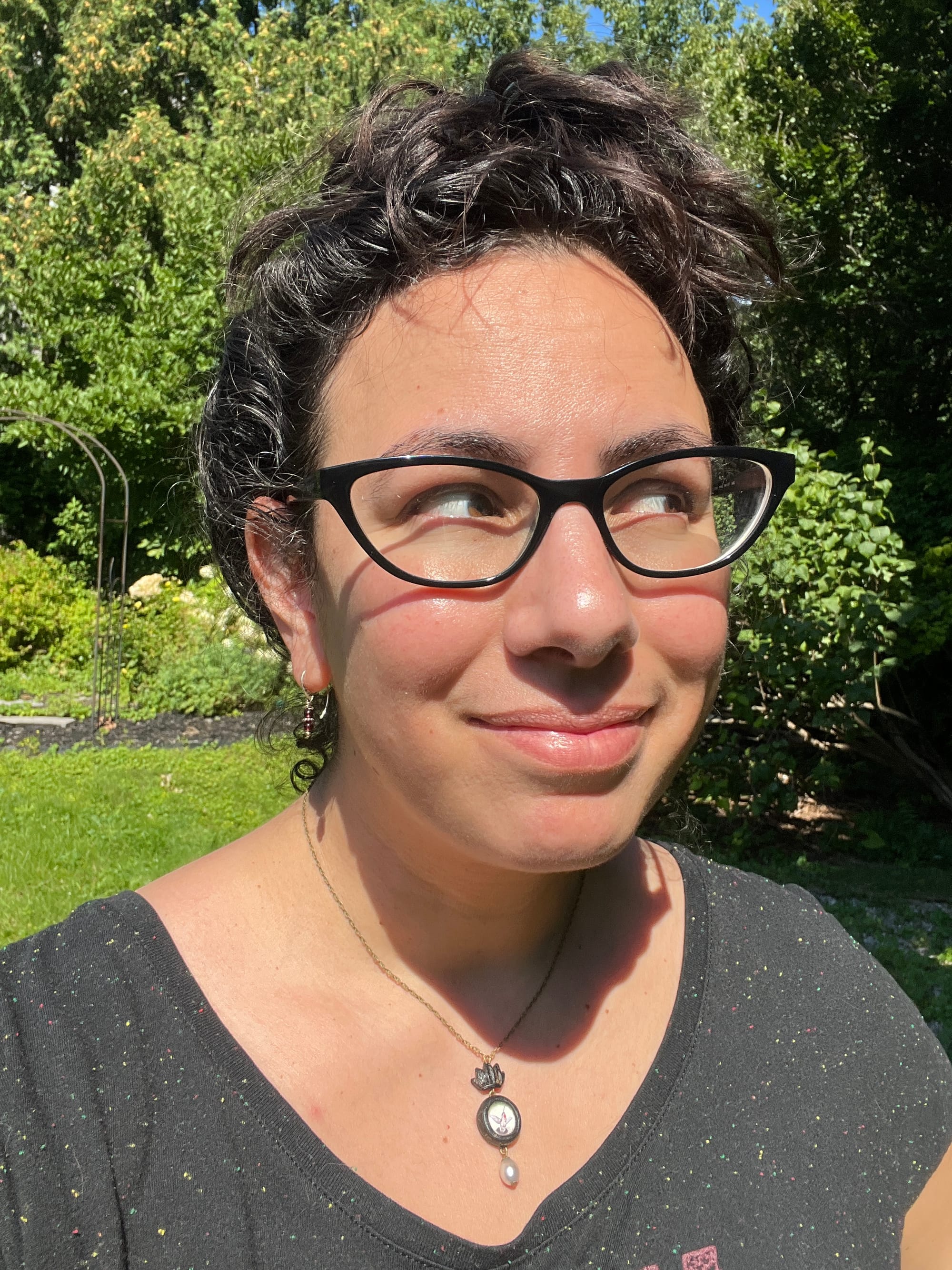 Sunny garden selfie in which I have my long dark wavy hair pinned up in a loose messy way that makes it look a bit like I have a pixie cut. My eyes are looking off to the side behind black-rimmed cat’s eye glasses and I’m smiling close-mouthed while wearing a loose-fitting Hadestown t-shirt, tiny garnet earrings, and a parrish relics necklace depicting a tiny painting of a hummingbird under glass with a pearl drop beneath.