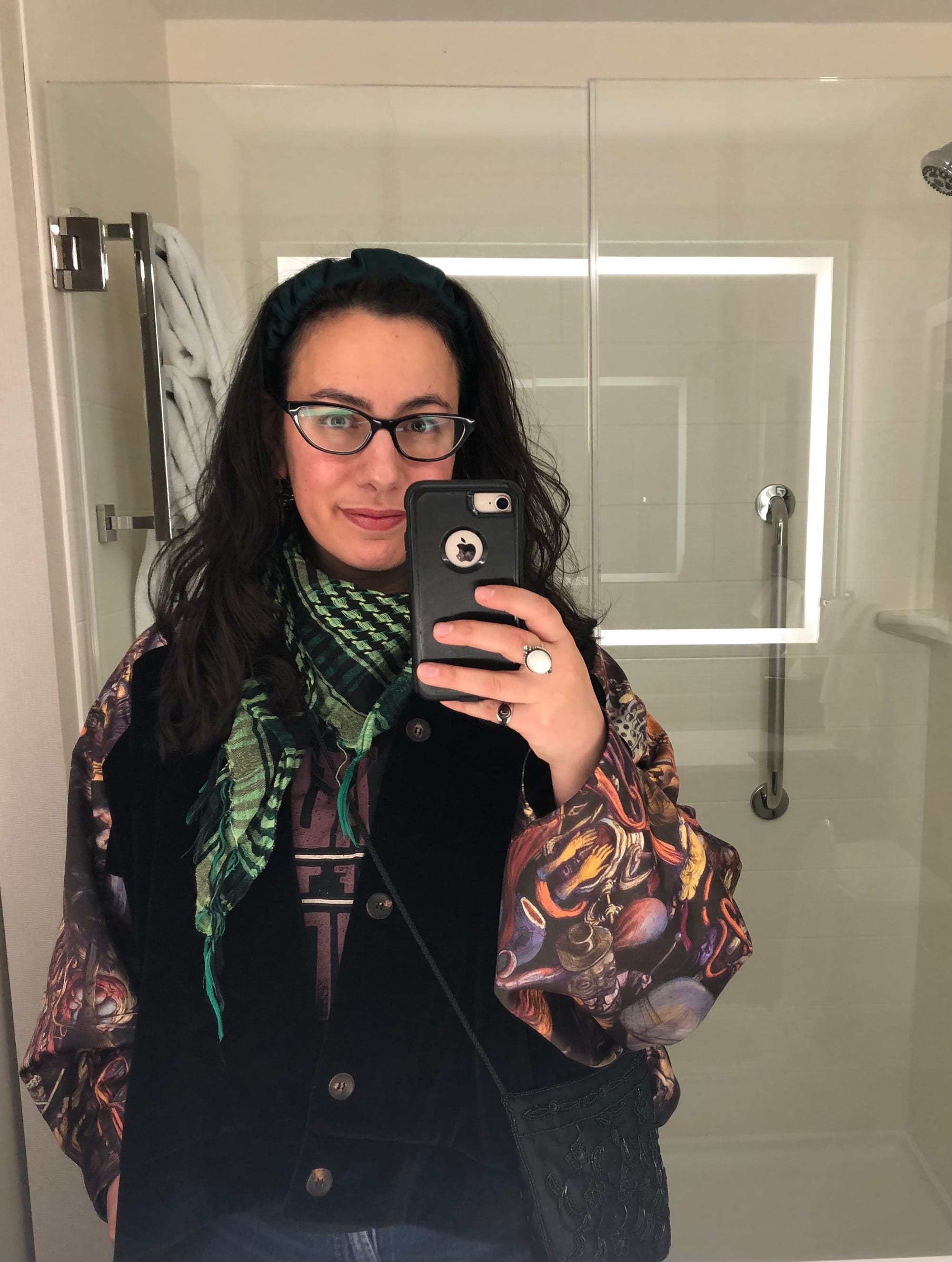 Mirror selfie in a hotel bathroom in which I'm wearing my dark wavy brown hair down around my shoulders, a green keffiyeh over a Hadestown t-shirt underneath a kimono-style jacket with art on it from the video game Disco Elysium, in chaotic almost Bosch-like figures and cityscape colours of yellow, orange, purple, against black. 