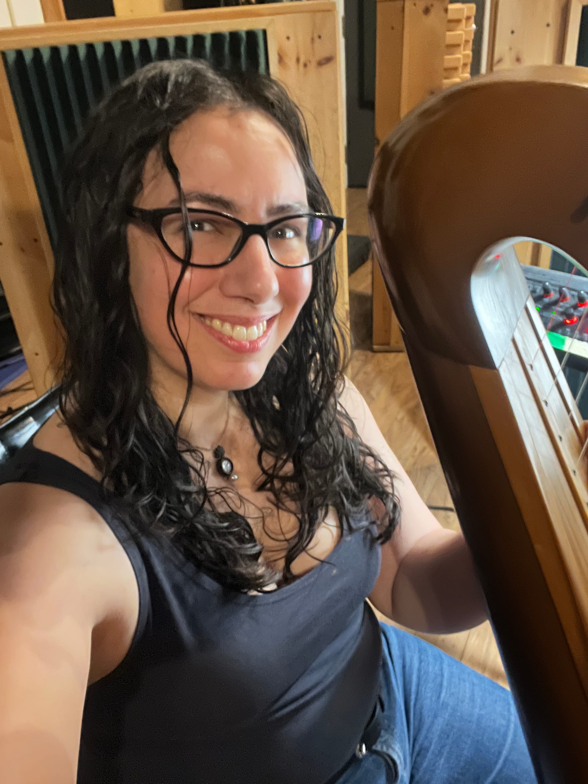 Selfie in which I'm seated at my harp, smiling at camera with my hair down in ringleting waves, wearing a grey tank top and jeans.