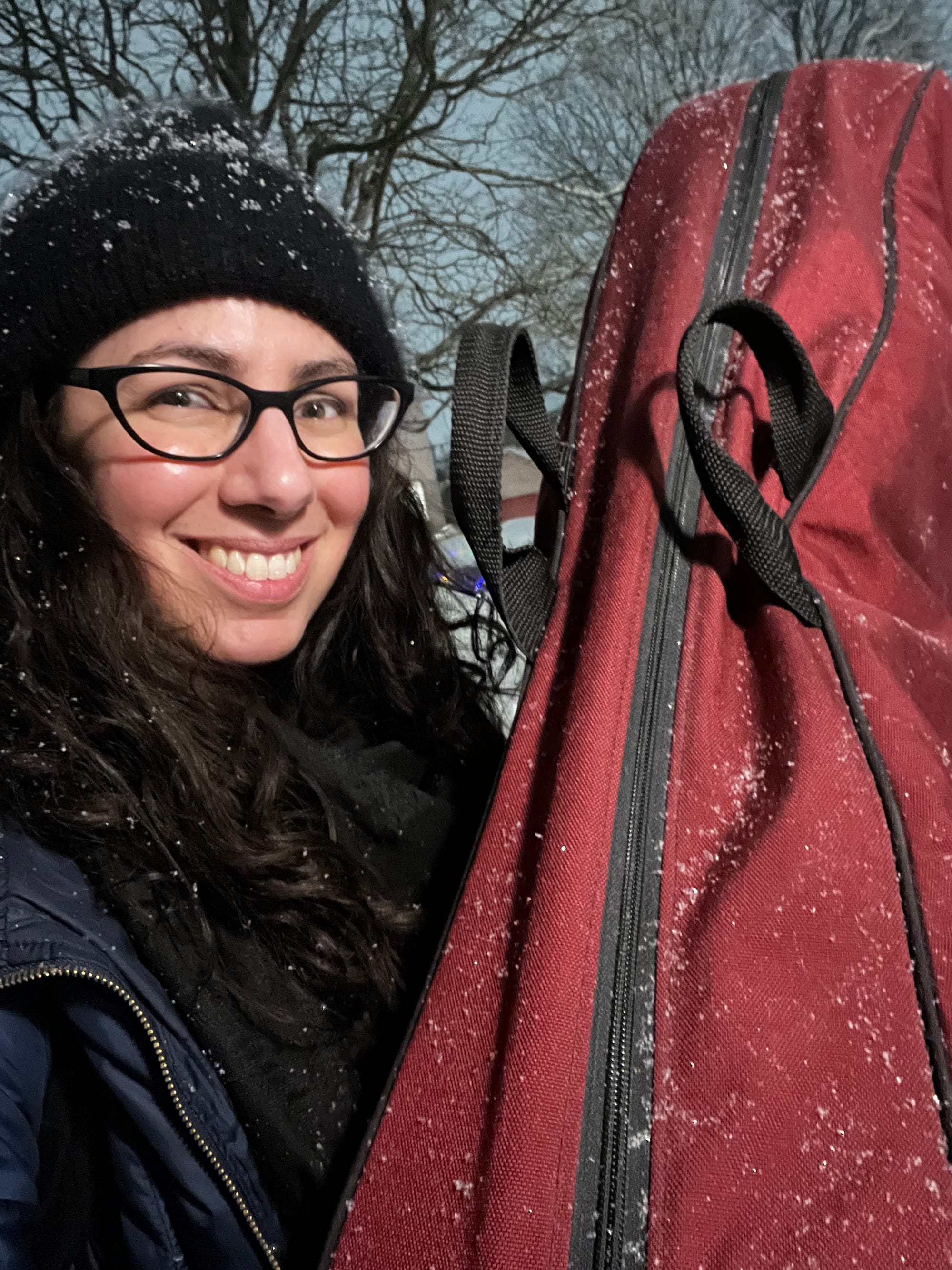 Selfie in which I'm grinning while holding the padded canvas bag containing my harp, slightly taller than I am, while snow falls on us both.