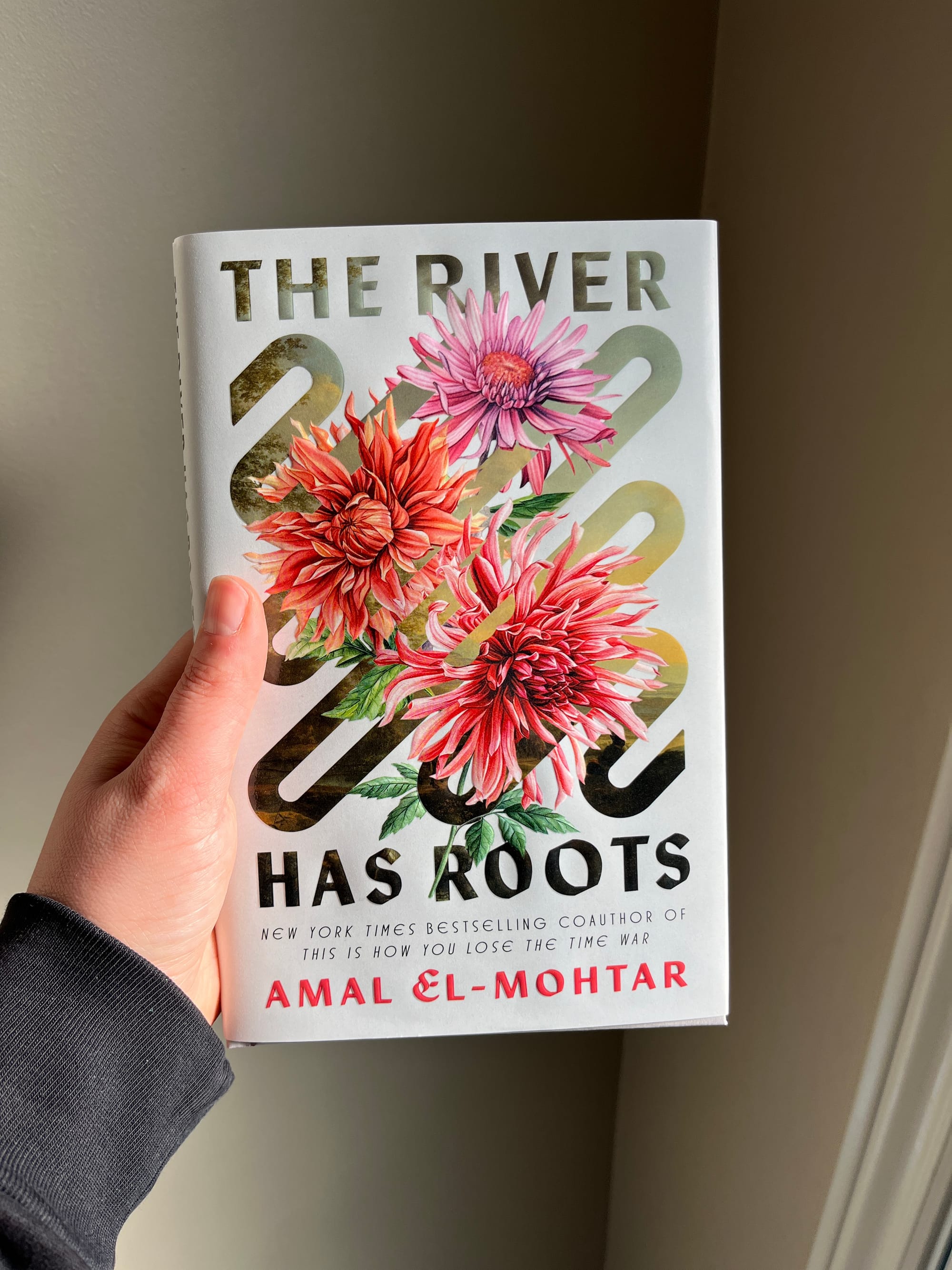 My left hand holding up the finished hardcover of THE RIVER HAS ROOTS. On a pale cover, a squiggly graphic line moving from the top left corner of the book down to the bottom right corner is a portal into a soft, romantic landscape of rivers and hills; bursting out between the squiggle’s curves are bright bold dahlias in warm hues. At the top of the image are the words “The River” and at the bottom are the words “Has Roots”; below those are “New York Times Bestselling author Amal El-Mohtar.”