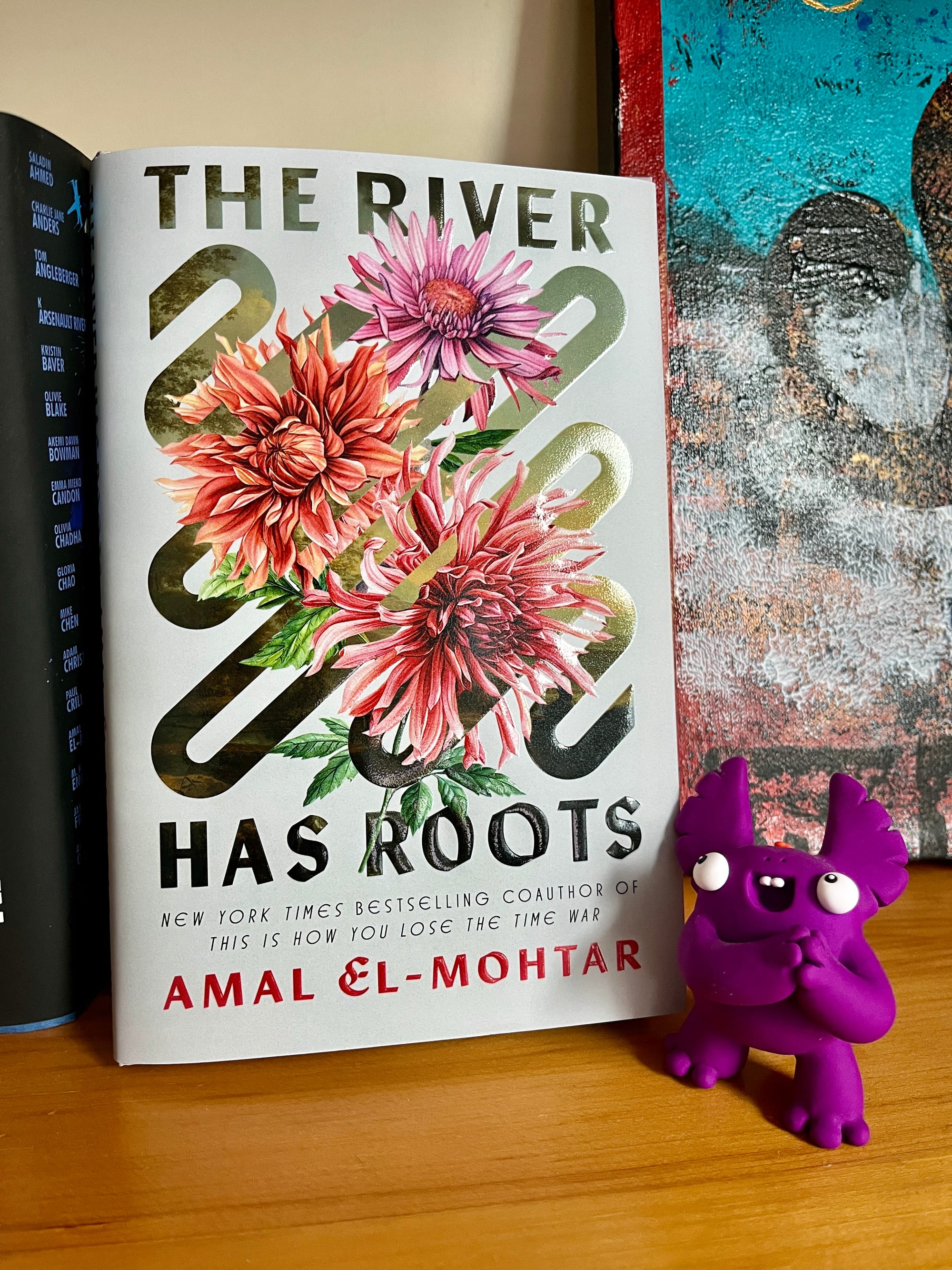 A hardcover copy of THE RIVER HAS ROOTS on a bookshelf, being admired by a small cartoonish sculpture of a wide-eyed purple beast clasping its little hands and grinning a buck-toothed grin.