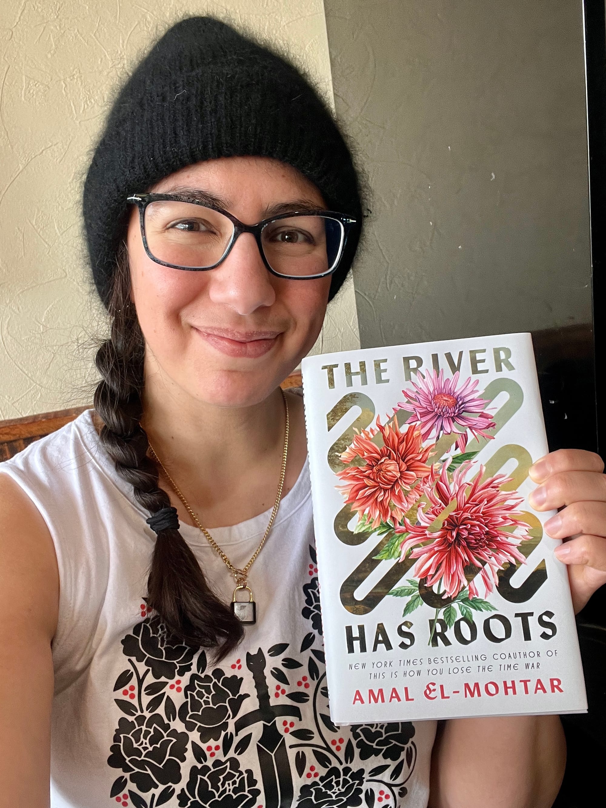 Selfie in which I'm wearing a fuzzy black beanie, wide square-ish glasses, my dark hair in a side braid over a white tank by Yoshi Yoshitano printed with black roses surrounding a sword and embellished with clusters of red berries, and a small black padlock necklace. I'm holding up a copy of THE RIVER HAS ROOTS and smiling.