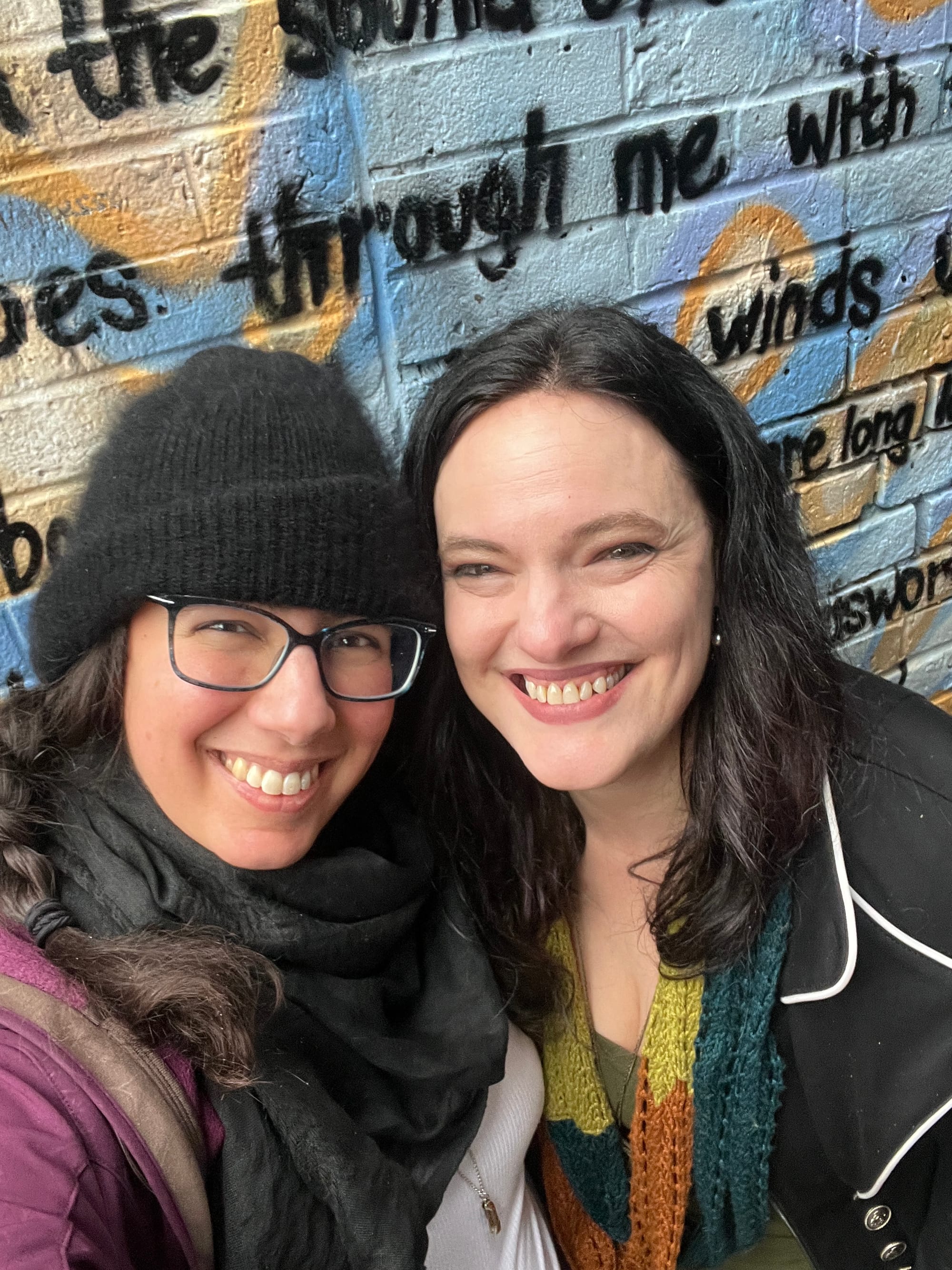 Selfie in which Cat and I are smiling at the camera; my dark hair's braided to the side and I'm wearing a fuzzy black beanie and an all-black keffiyeh with a dark purple jacket over a white tank top, and Cat's wearing her dark hair down with a colourful hand-knitted scarf she made herself over an olive green dress with a black military-style jacket with white popping on the lapels and silver buttons. Behind us is a mural on a brick wall of a poem, of which only a few words are visible ("through me with ... winds")