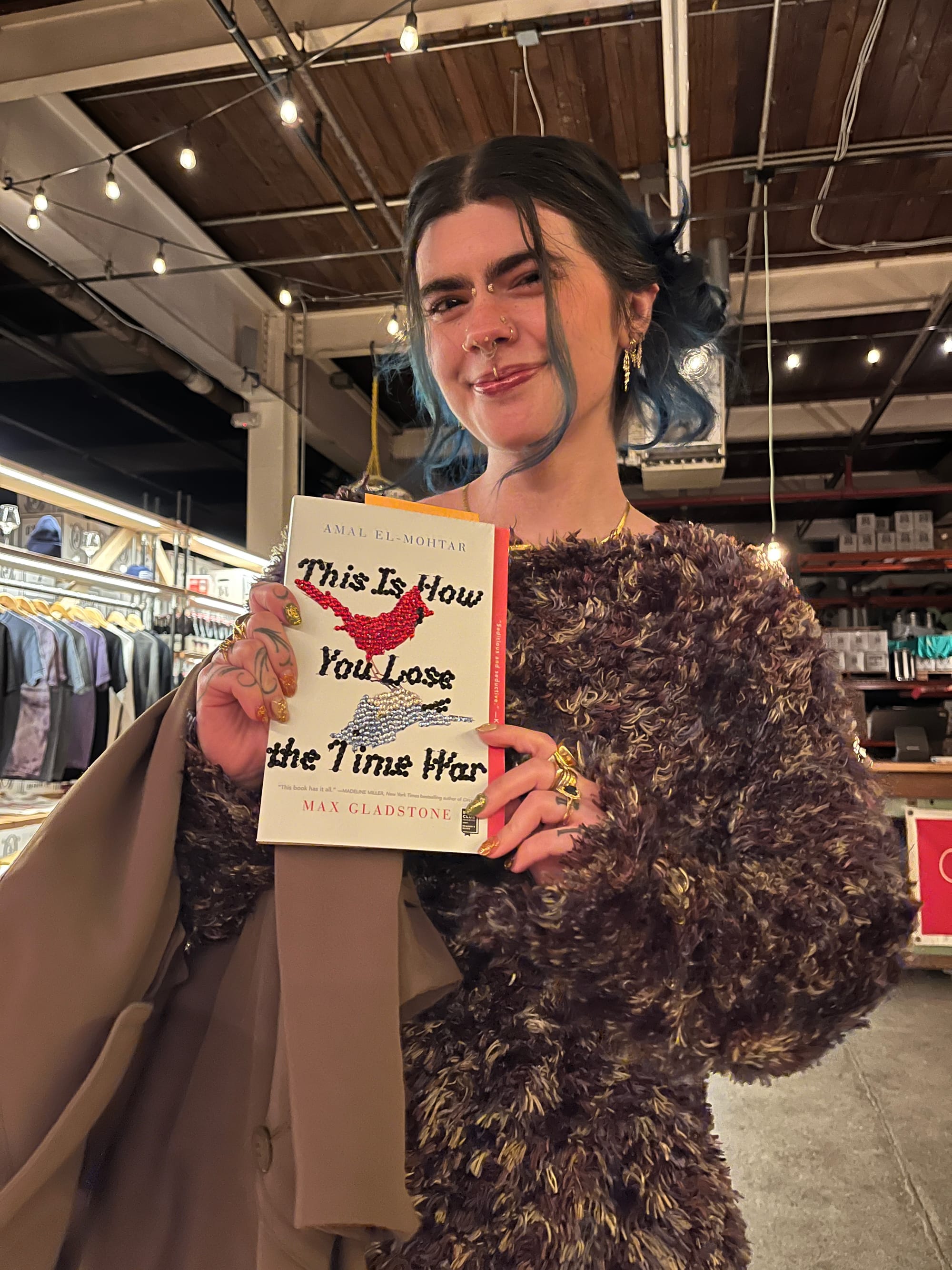 A reader with fabulous personal style (fluffy jumper, gorgeous nails and rings, blue dip-dyed hair and a lot of earrings and facial piercings) holding up a copy of This Is How You Lose the Time War having bedazzled the cover with sequins. 