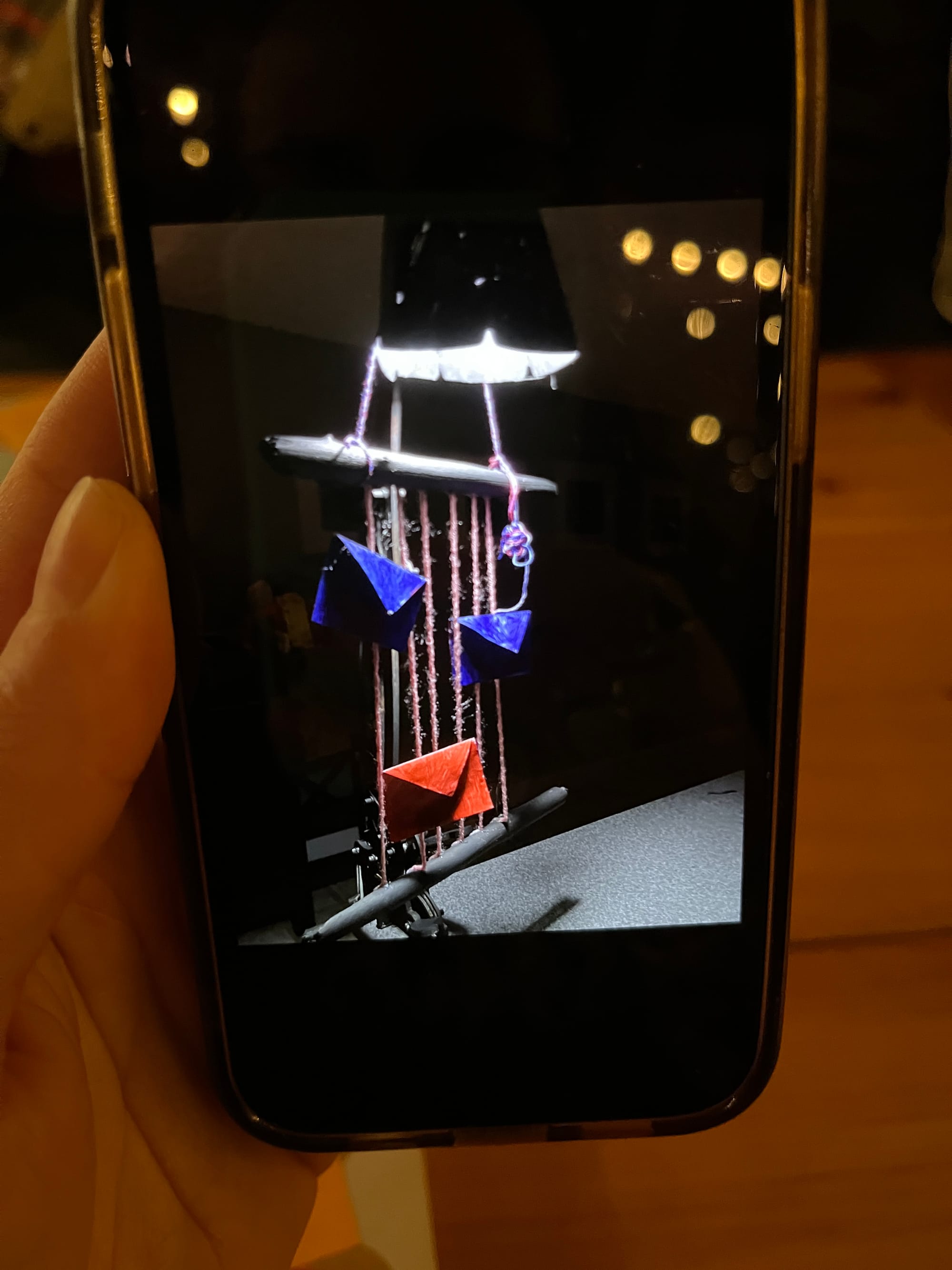 My hand is holding a phone on which is displayed a photo of handmade art object: a lamp suspended above a makeshift loom, from which dangle three painted envelopes, suggesting letters, one in red and two in blue.