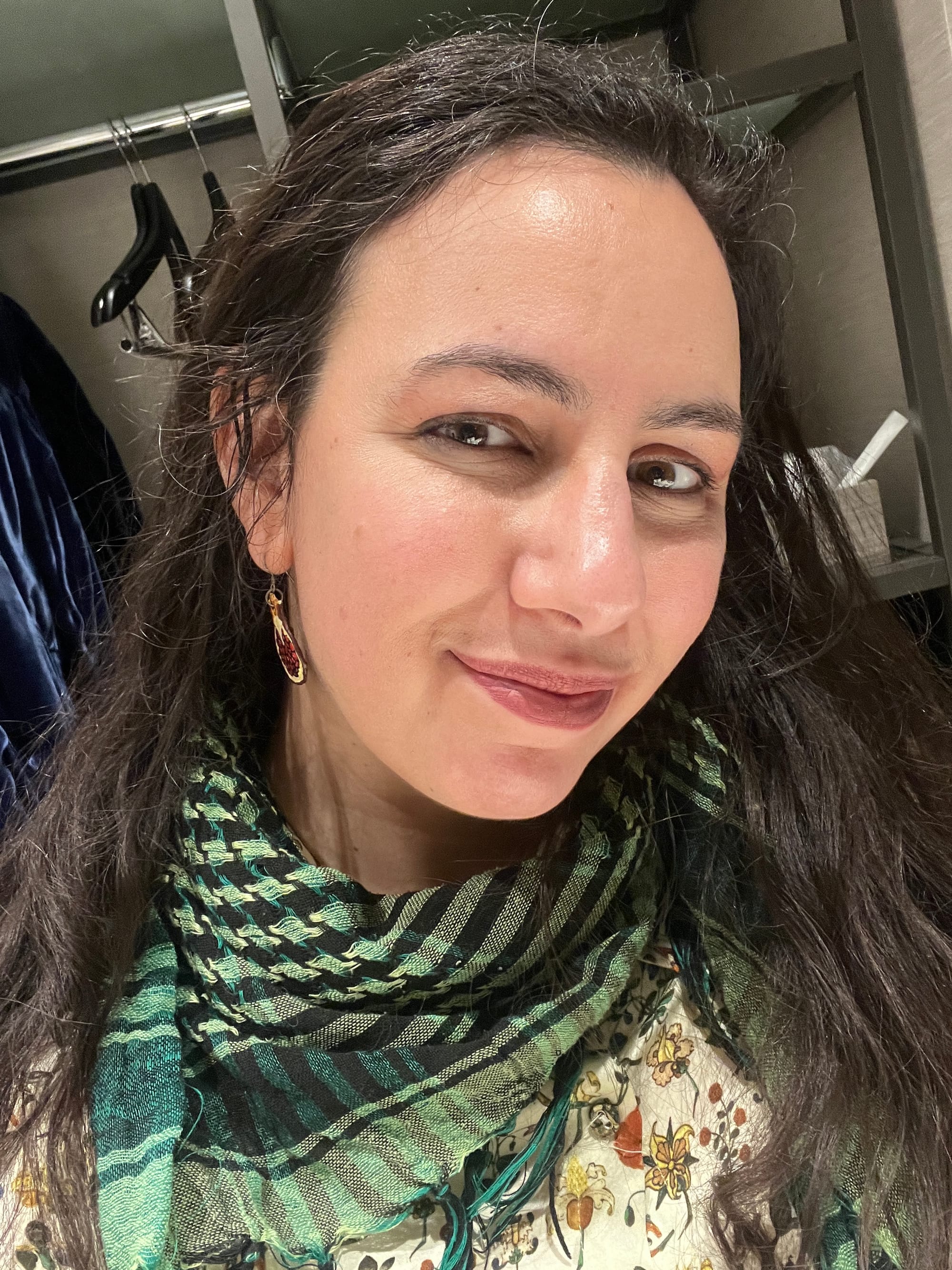 A post-event hotel room selfie in which I'm wearing my long wavy dark hair down, with a green and black keffiyeh over a Samantha Pleet blouse in a medieval illumination print featuring flowers and fruits. The bad lighting is being heroically combatted by foundation, eyeshadow, eyeliner and lipstick, which I rarely put on myself but am always happy to have skilled people like Cat Valente put on me.