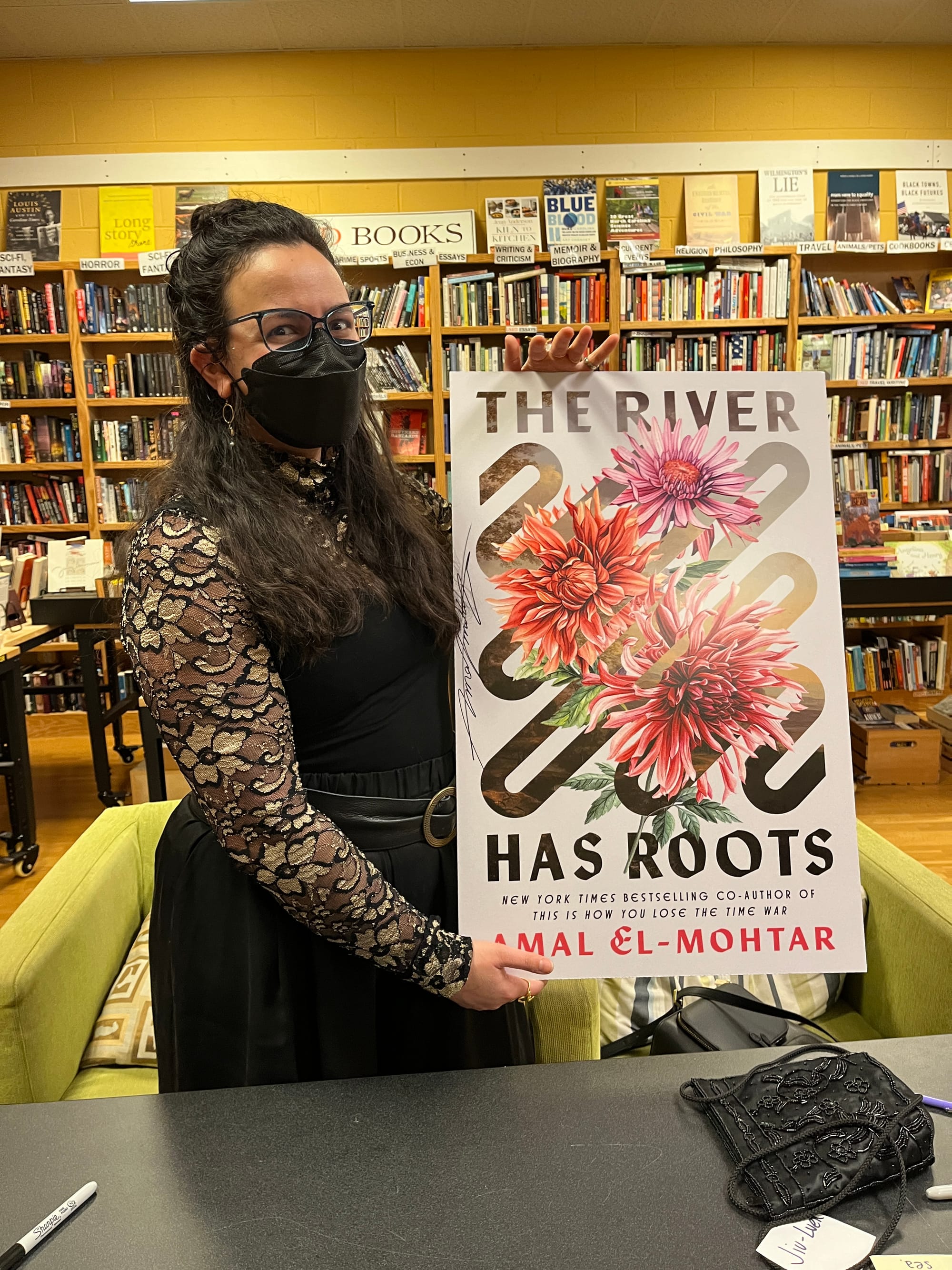 Photo of me holding up a giant version of the cover to THE RIVER HAS ROOTS. I'm wearing my dark wavy hair half-up, half-down, a black mask, and a lacy floral top with gold accents layered under a black tank top with swishy wide-legged black trousers and a wide leatherish belt with a round brass buckle. In the background are shelves full of books.