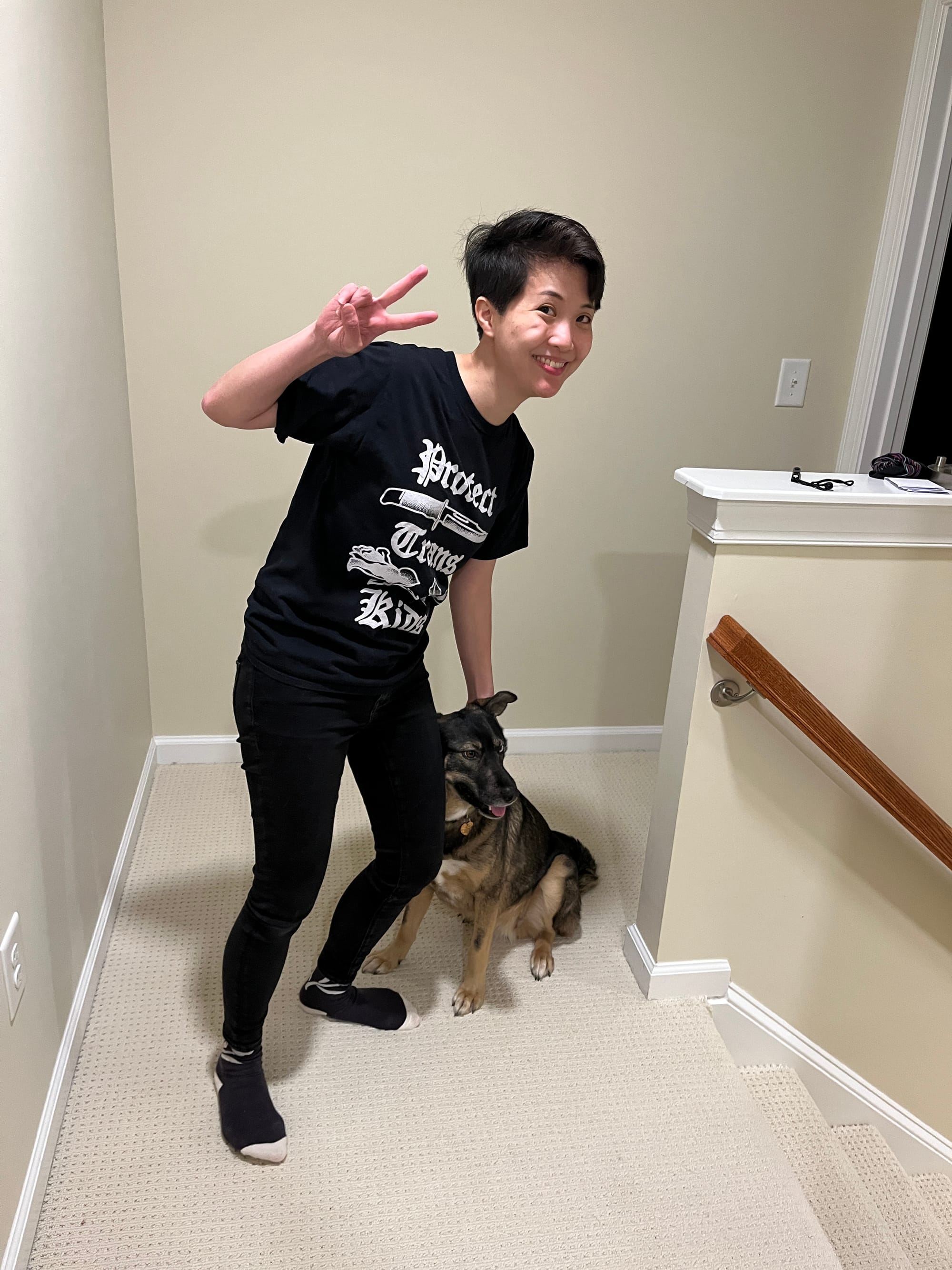 Alyssa Wong, a brilliant author, wearing black trousers and a black t-shirt that reads "Protect Trans Kids" in a gothic font illustrated with a knife and rose. They're throwing a peace sign while petting an adorable dog that looks like a tiny German shepherd.