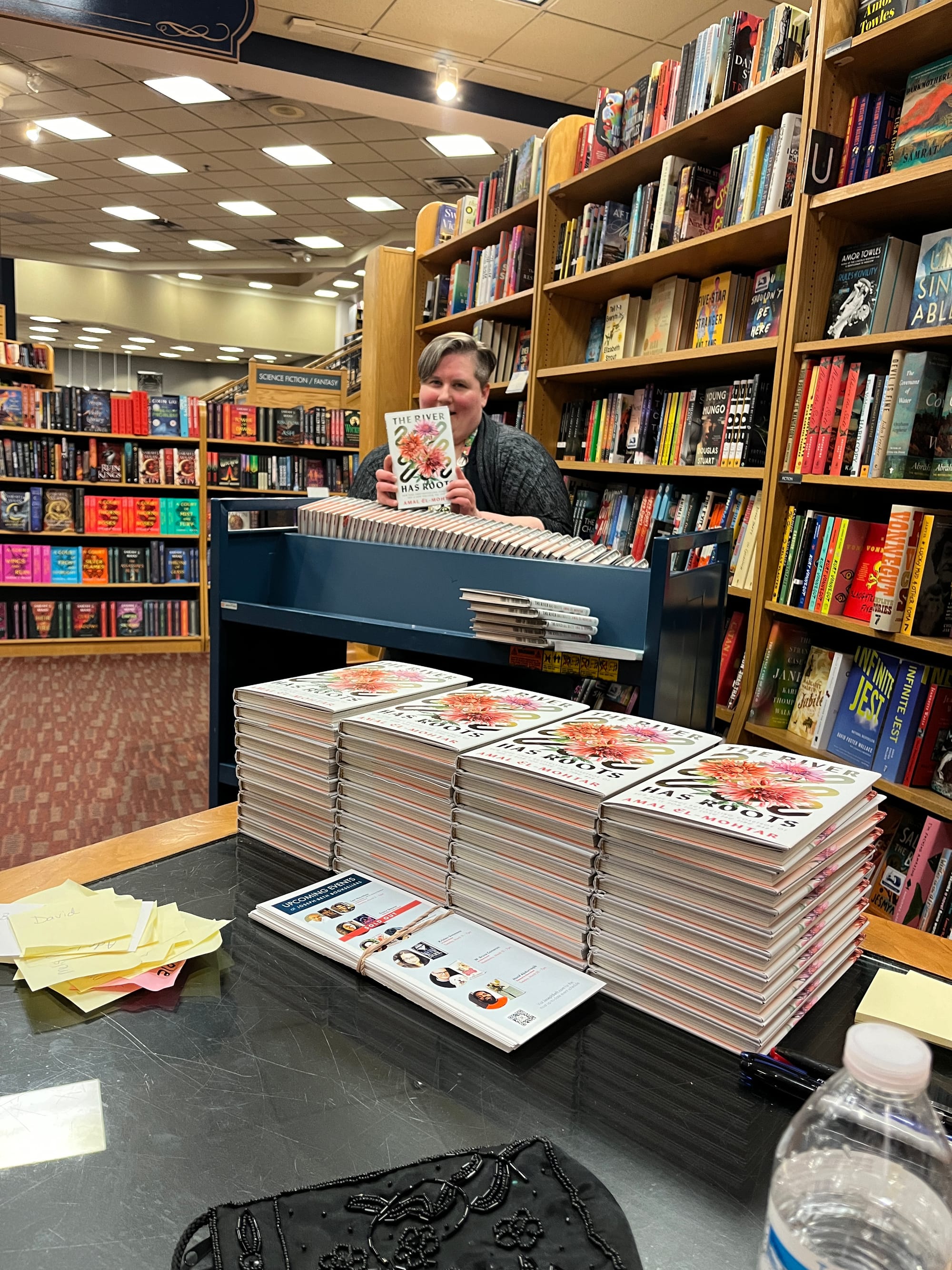 A photo taken at Joseph-Beth Booksellers in Cincinnati. Four stacks of ten copies each of THE RIVER HAS ROOTS in the centre, while behind them is a book cart with a whole shelf full of more copies, behind which is a bookseller holding up one more copy partially obscuring their face.