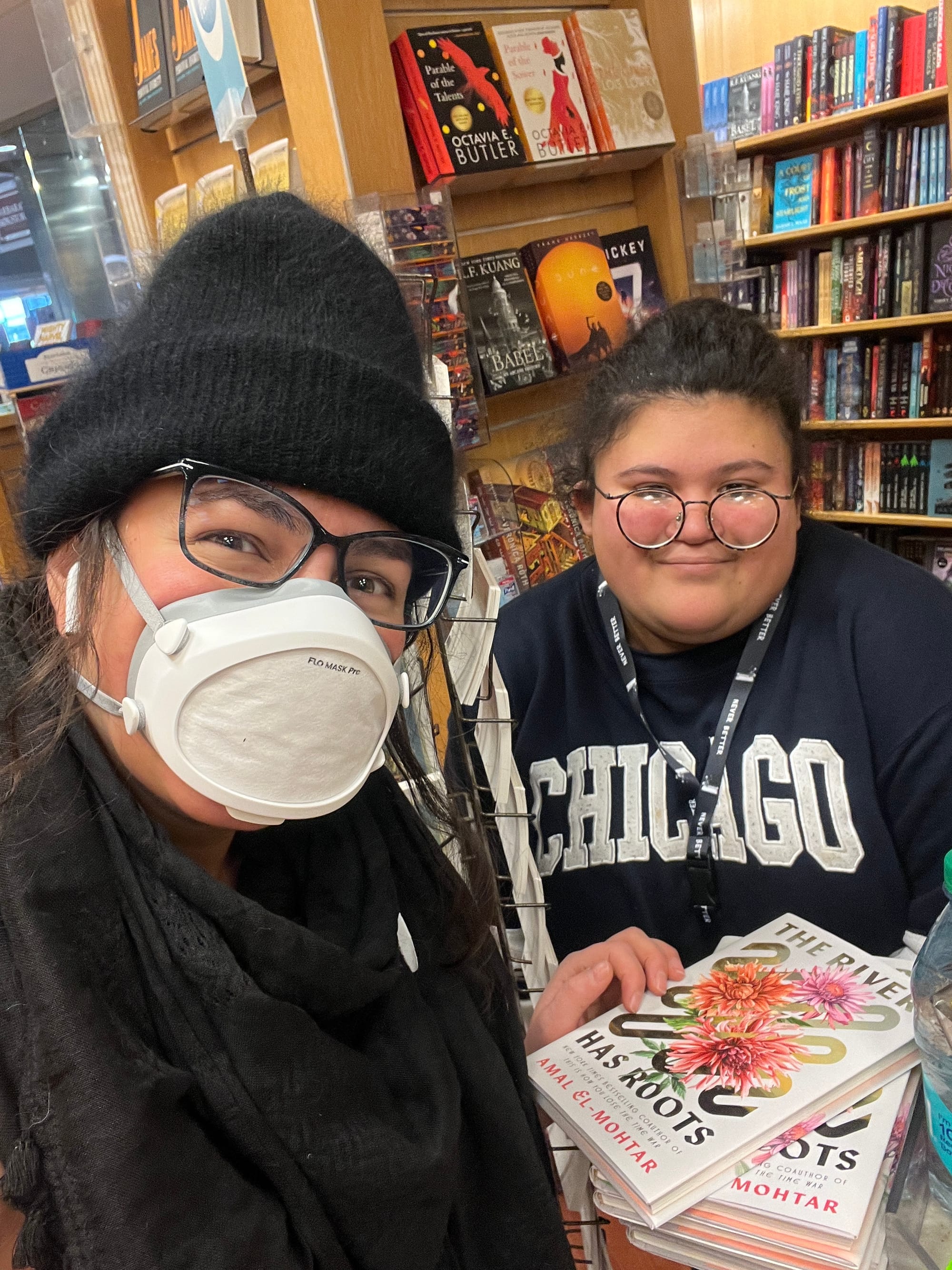 Selfie in which I’m wearing a flo mask, black fuzzy beanie and black-on-black keffiyeh next to a bookseller wearing a shirt that reads CHICAGO. Between us is a stack of signed copies of my book.