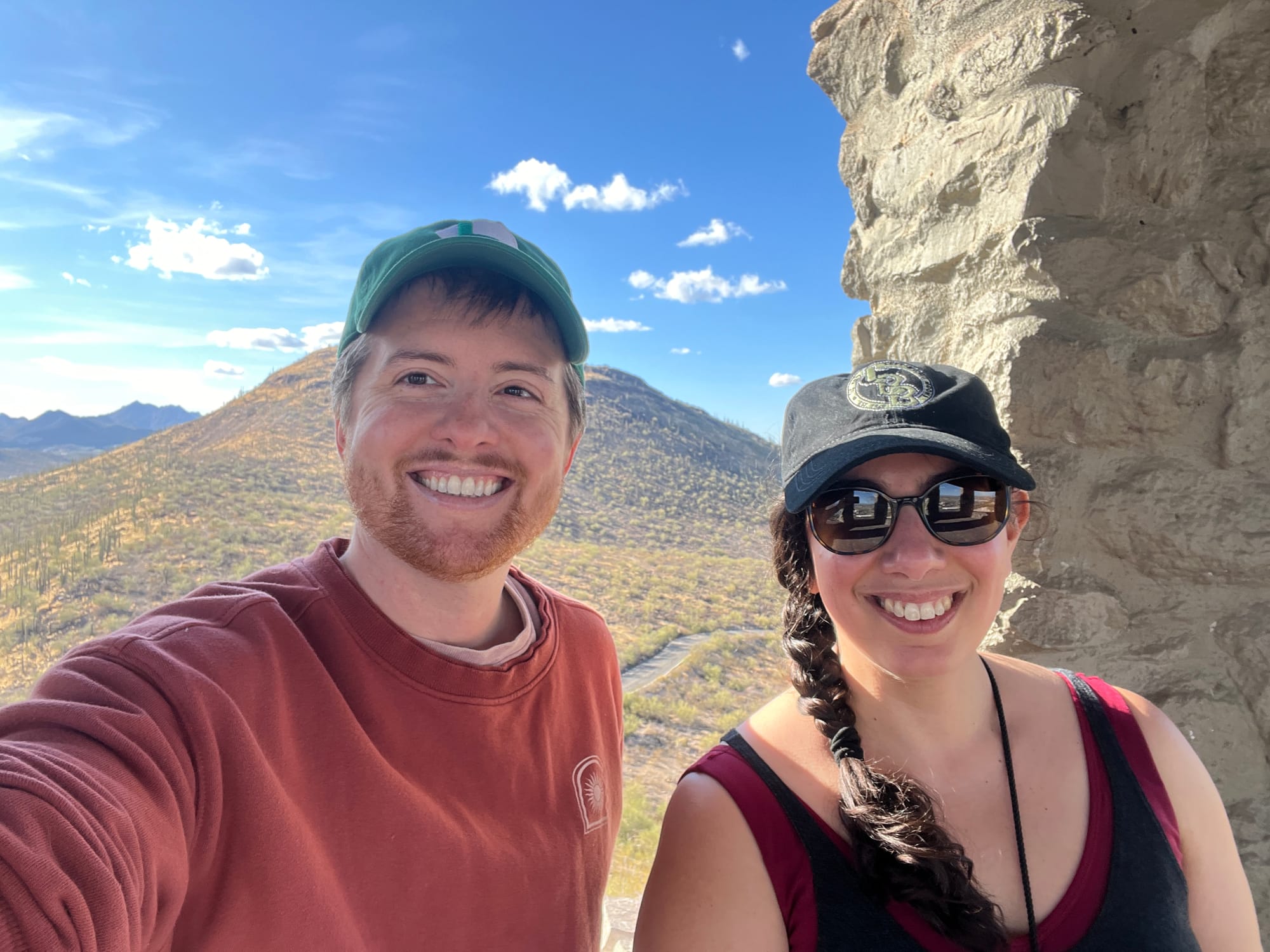 A selfie in which Danny -- a man with a ginger beard, red sweatshirt and green baseball cap -- and I (a woman with dark hair in a side braid under a black baseball cap, wearing sunglasses and a black tank top over a red sports bra) standing in front of a saguaro-covered mountain in the background, with a mortared stone pillar behind us to the right of the photo. The sky is very blue and bright, dotted with fluffy white clouds.